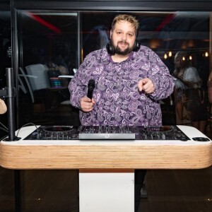 A DJ with headphones around his neck wearing a purple paisley shirt, holding a microphone in one hand and pointing forward while standing behind a mixing console.