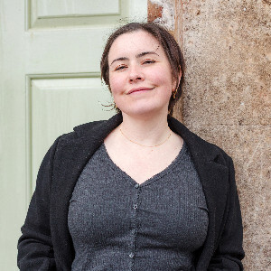 Katy Brothers, wedding photographer, smiling against a rustic wall—warm, approachable style. Book Katy Brothers Wedding Photography for natural, heartfelt wedding photos.