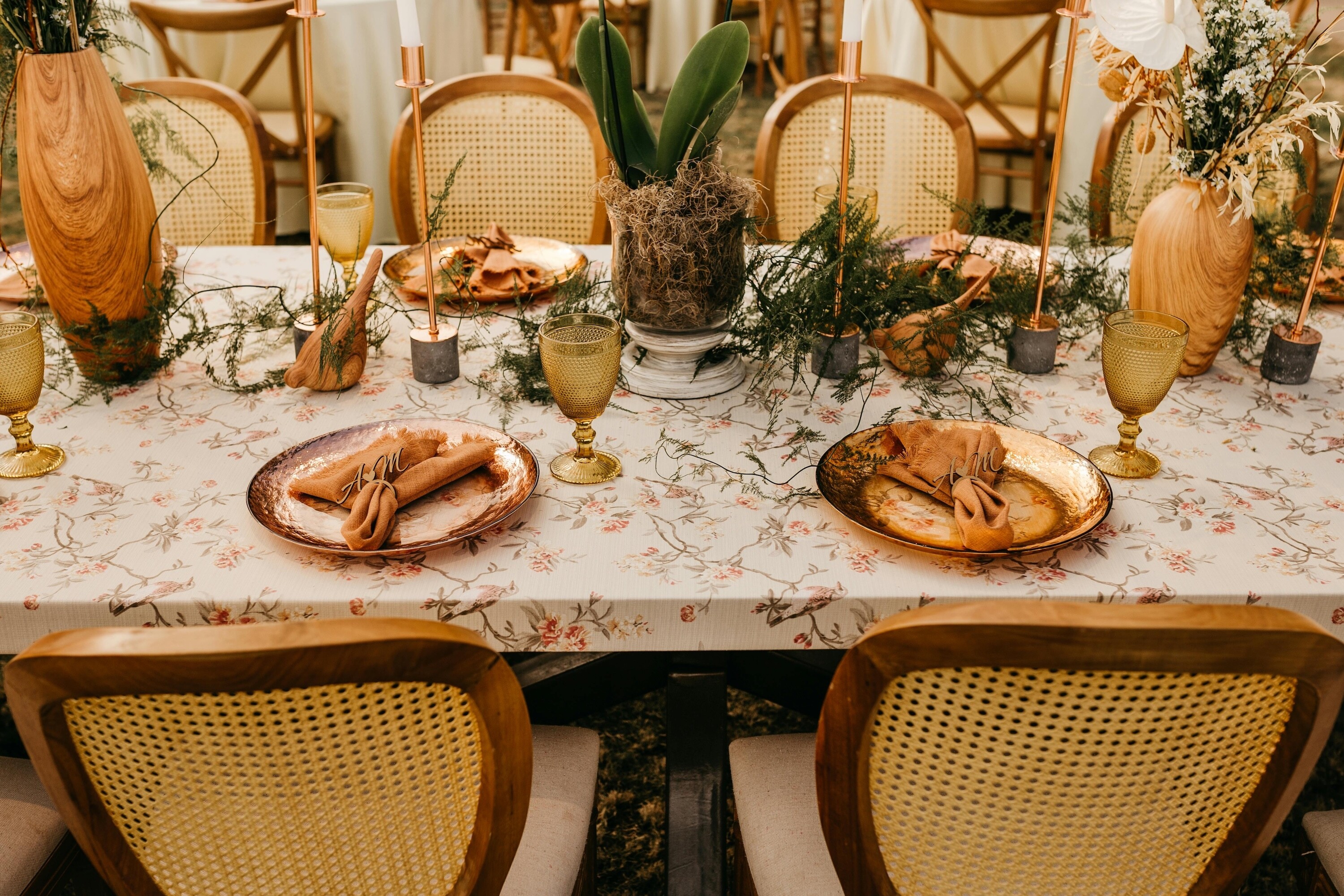 Rustic wedding tablescape with floral linen, gold plates, wooden chairs, candle holders and natural greenery styling.