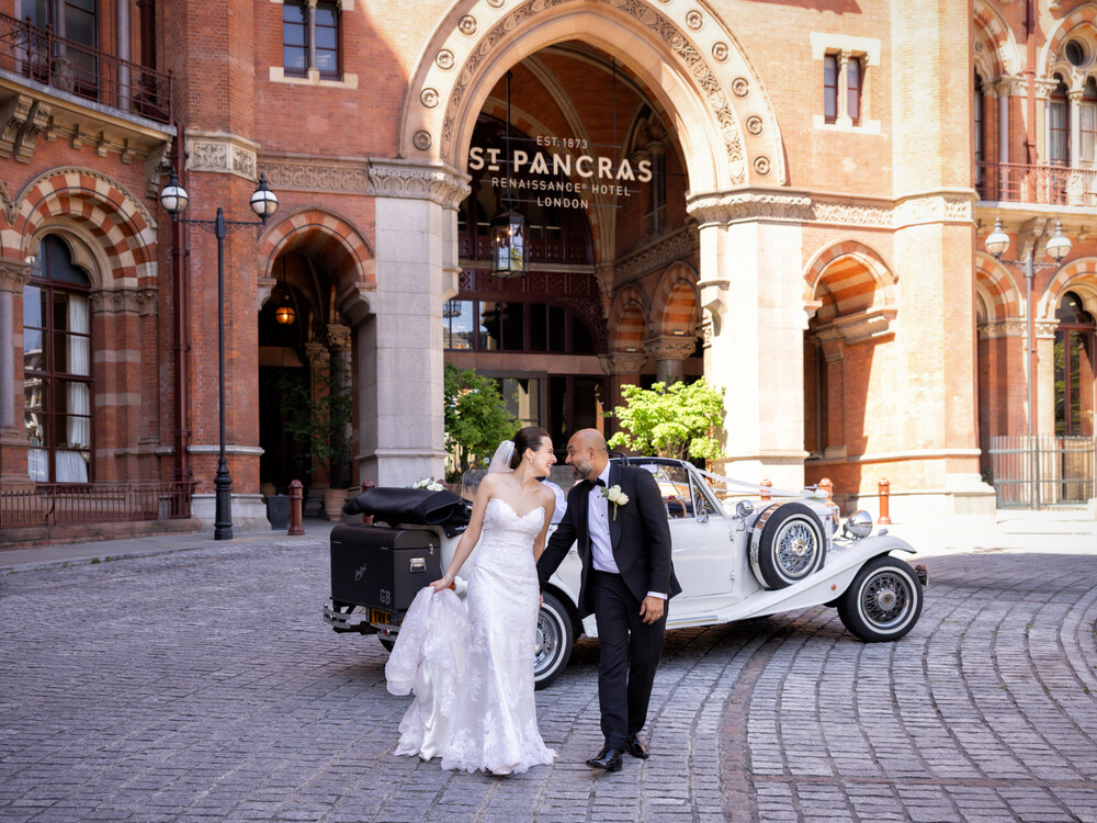 Bride and groom walking hand in hand outside St Pancras Renaissance Hotel in London, with a vintage wedding car parked behind them.