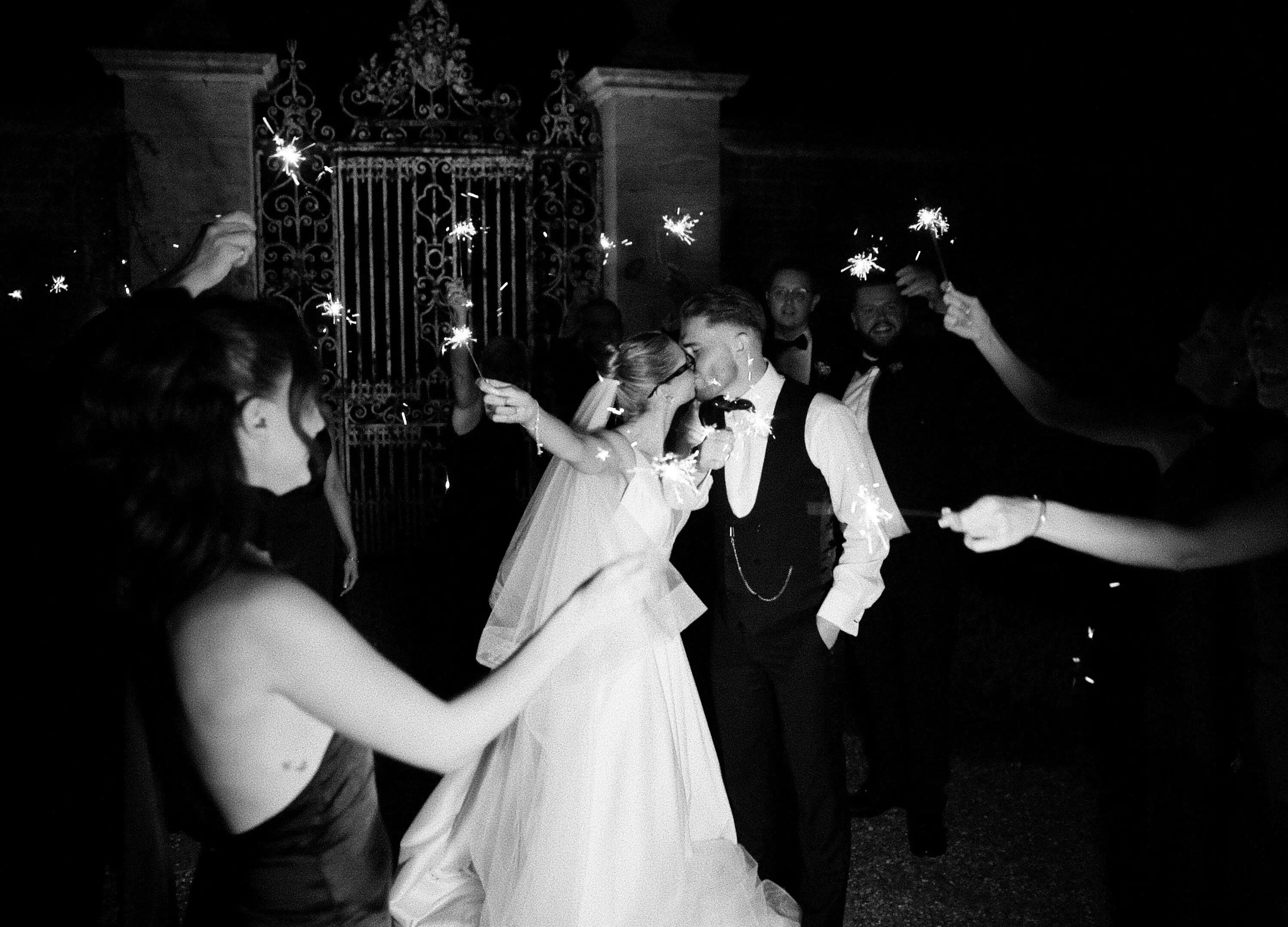 Black and white photo of a bride and groom kissing during a sparkler send-off, surrounded by guests holding sparklers outside an ornate gate at night.