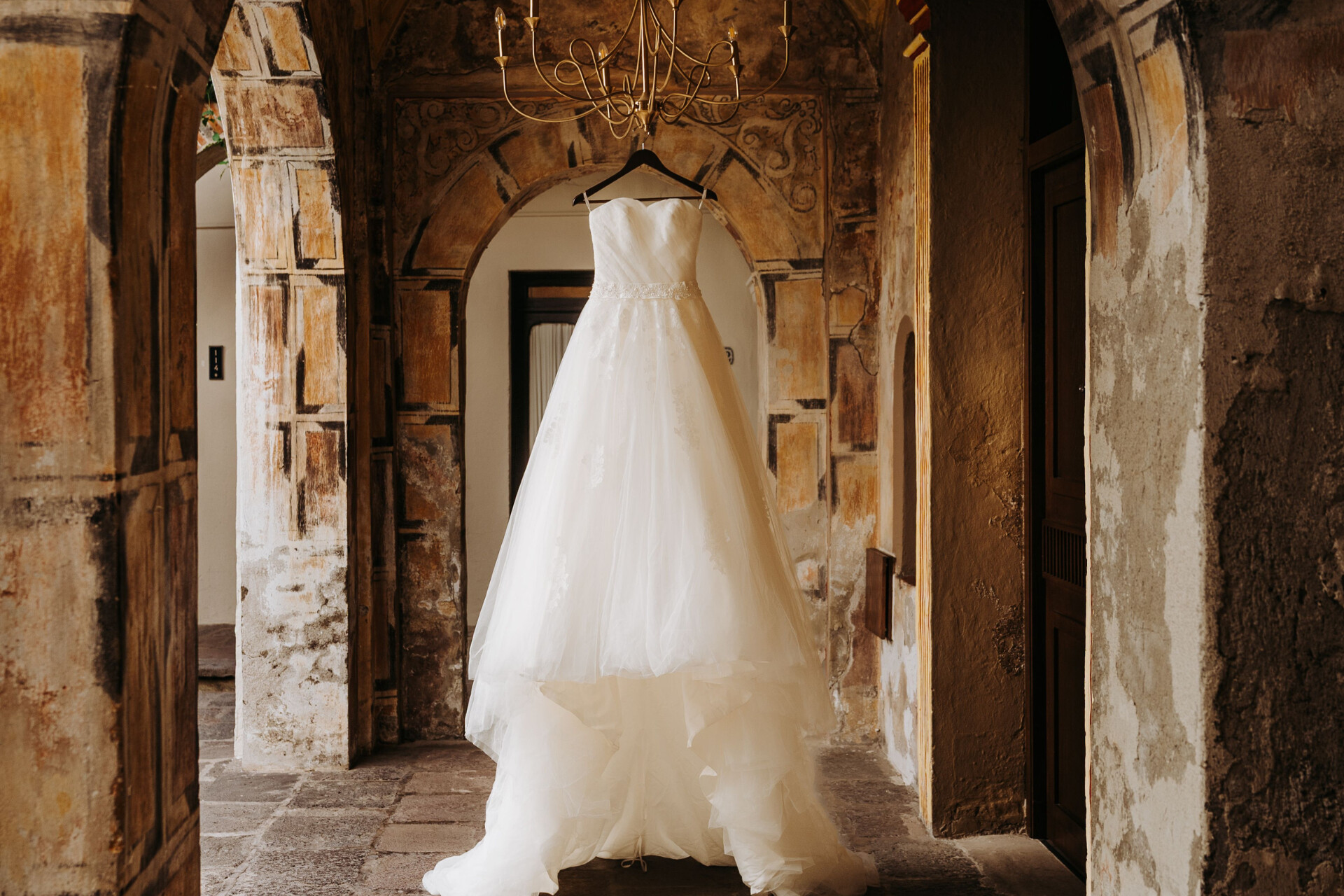 Elegant wedding dress displayed beneath a chandelier in a historic venue corridor.