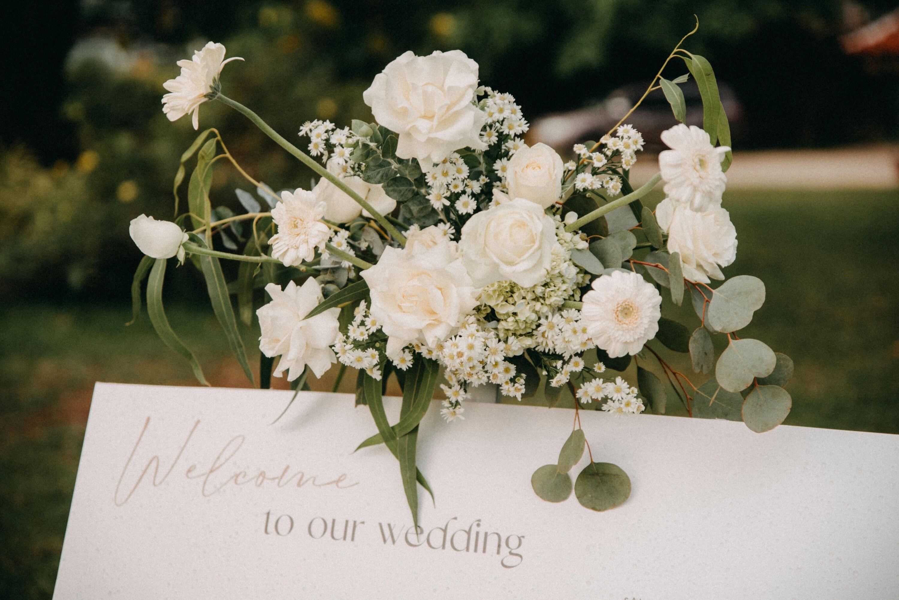 White wedding welcome sign decorated with white roses, daisies and eucalyptus flowers in an outdoor garden setting.