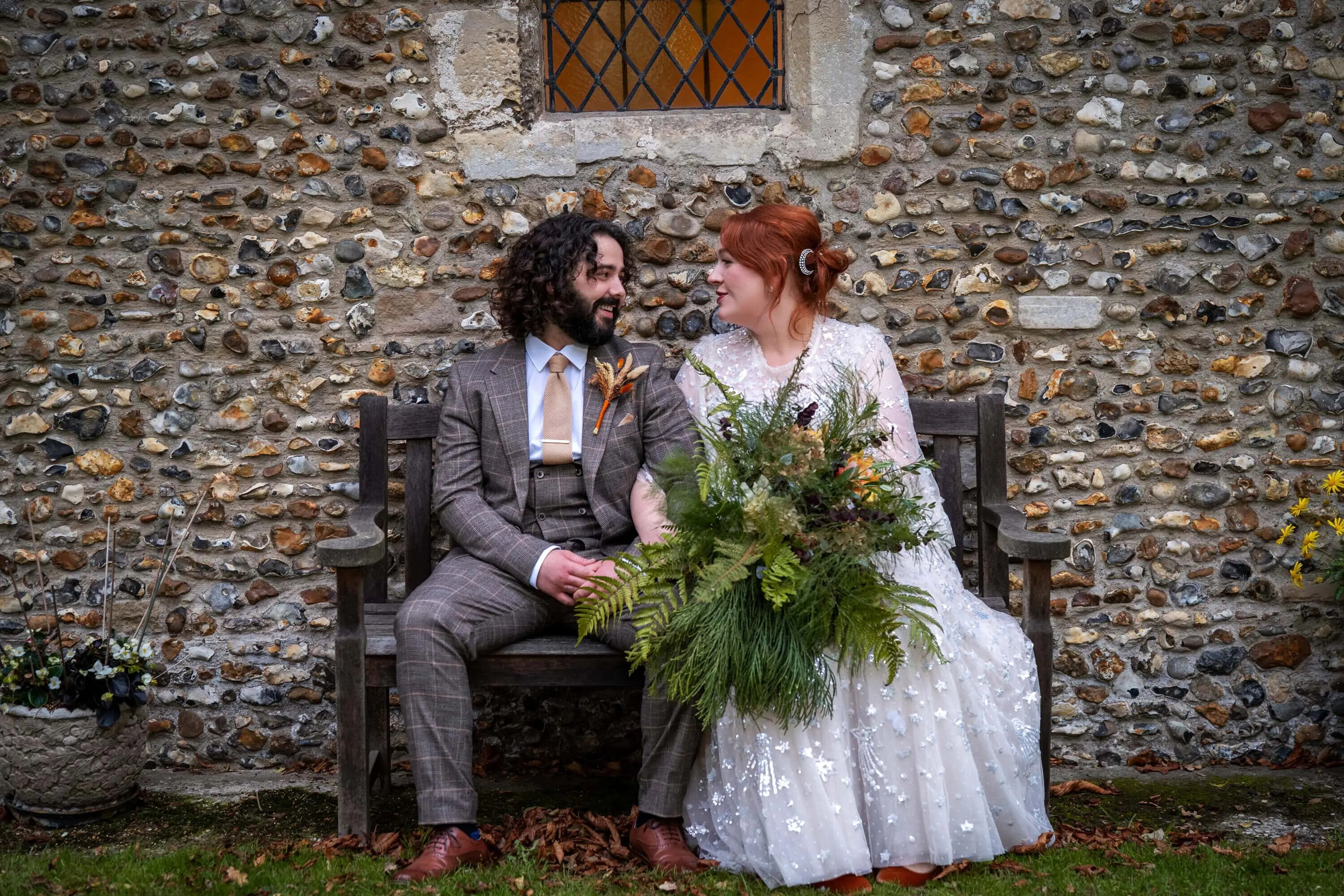Bride and groom sitting on a wooden bench outside a flint-stone church, smiling at each other on their wedding day, with the bride holding a large foliage bouquet.