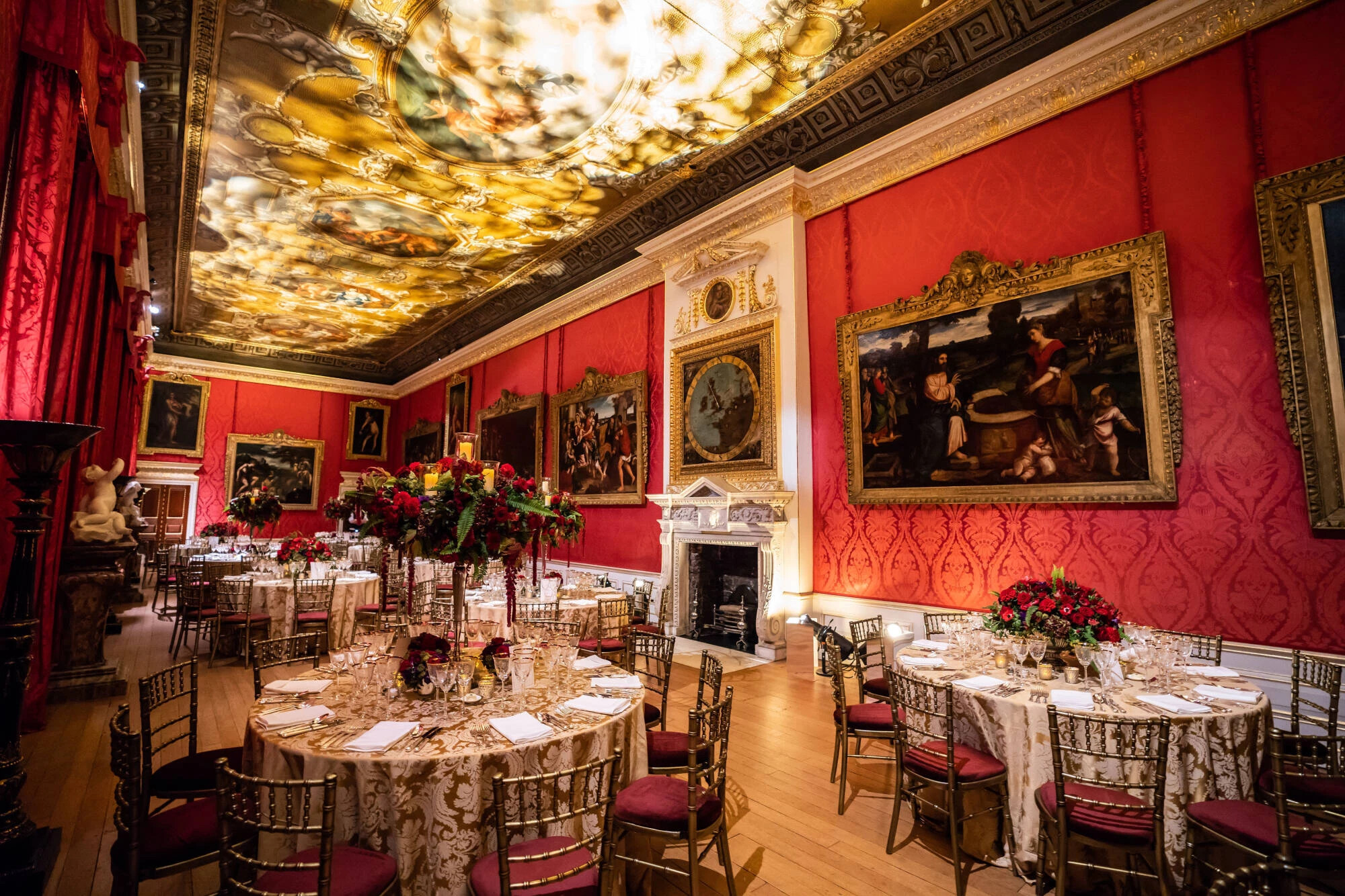 Interior of state room with red walls in Kensington Palace - space is set for a wedding breakfast with red floral displays in the centre of round tables.