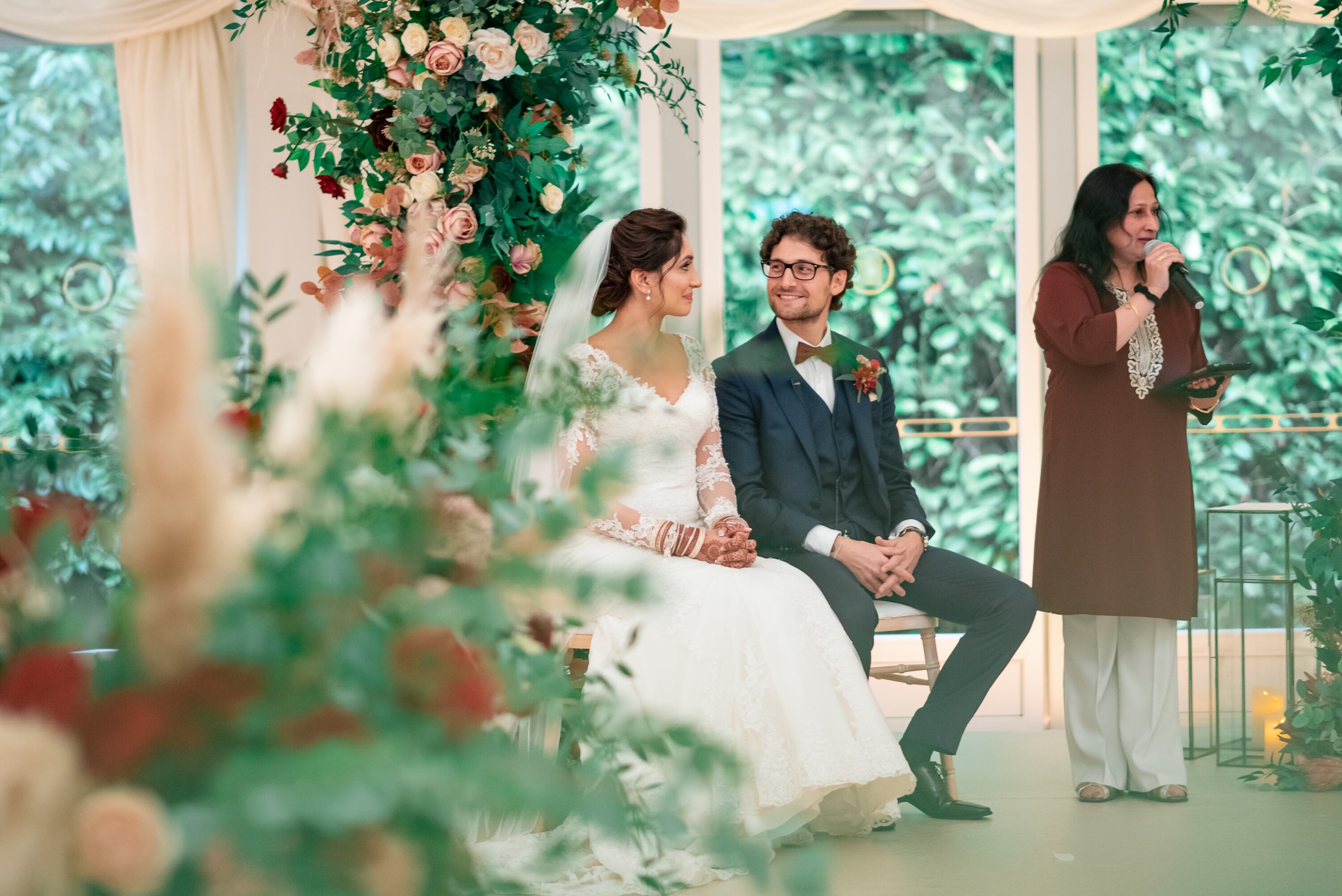 Sonal Dave, multicultural wedding celebrant, speaking during a wedding ceremony as the bride and groom sit beneath a floral arch.