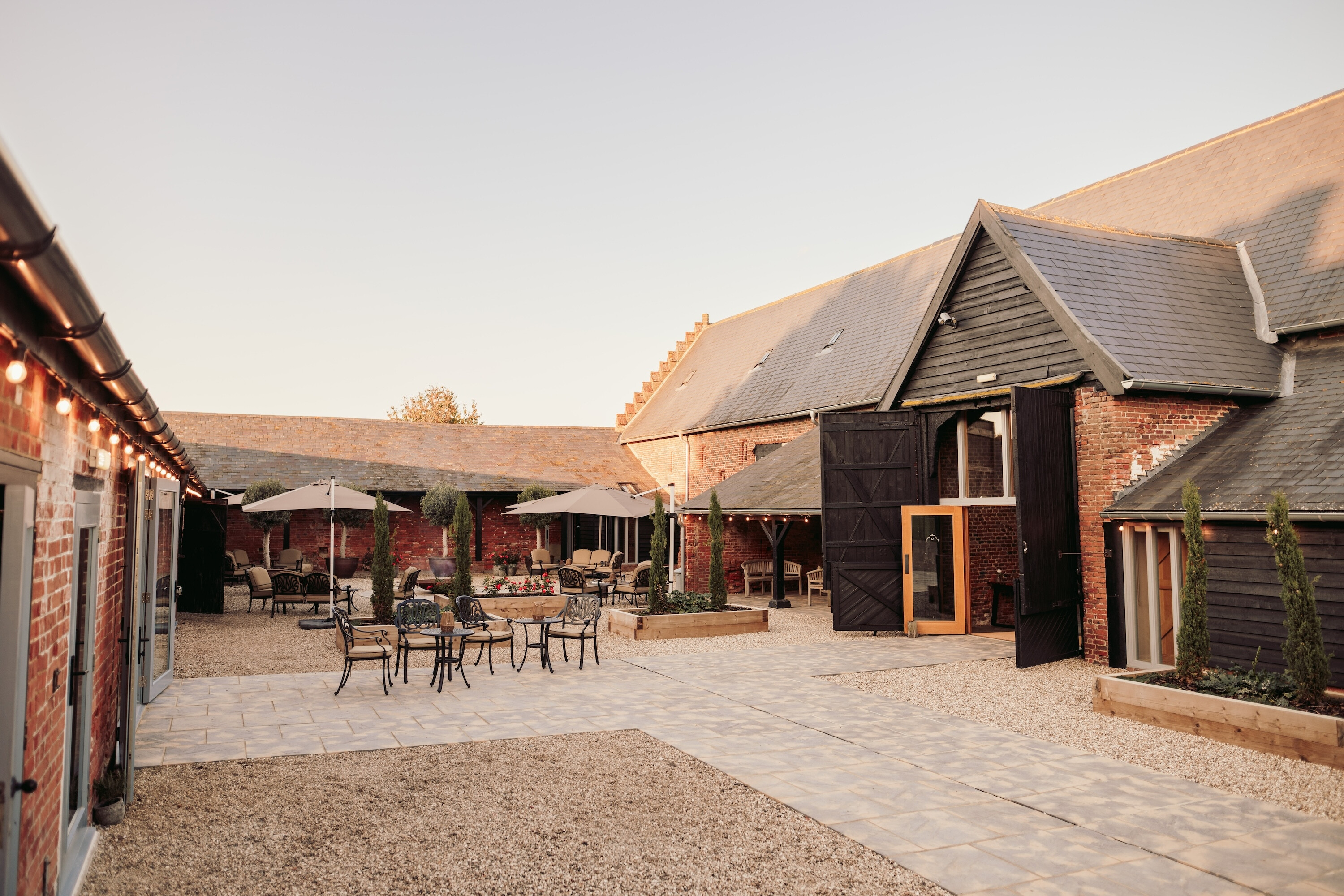 Courtyard at Copdock Hall with restored brick barns, outdoor seating, parasols and warm evening light in the Suffolk countryside.