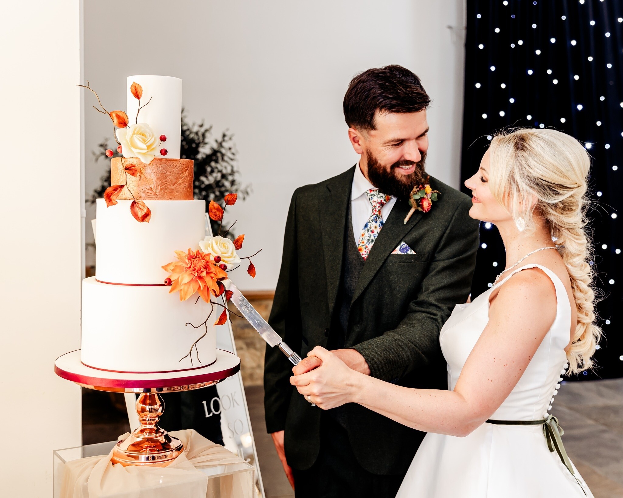 Bride and groom cutting a tall white tiered wedding cake by Cakes of Curiosity, decorated with copper detailing and autumn flowers on a rose-gold stand.