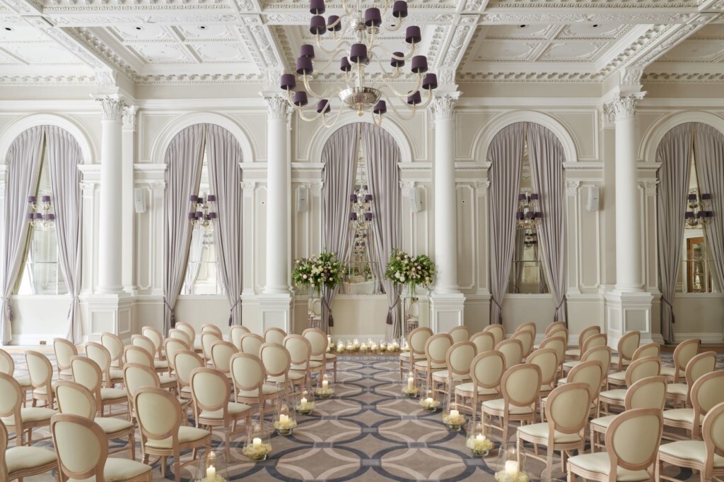 Elegant ceremony room at Corinthia London with tall columns, arched windows, draped curtains and rows of chairs set for a wedding ceremony.