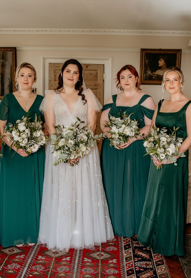Bride with bridesmaids in green dresses holding bouquets in a living room setting.