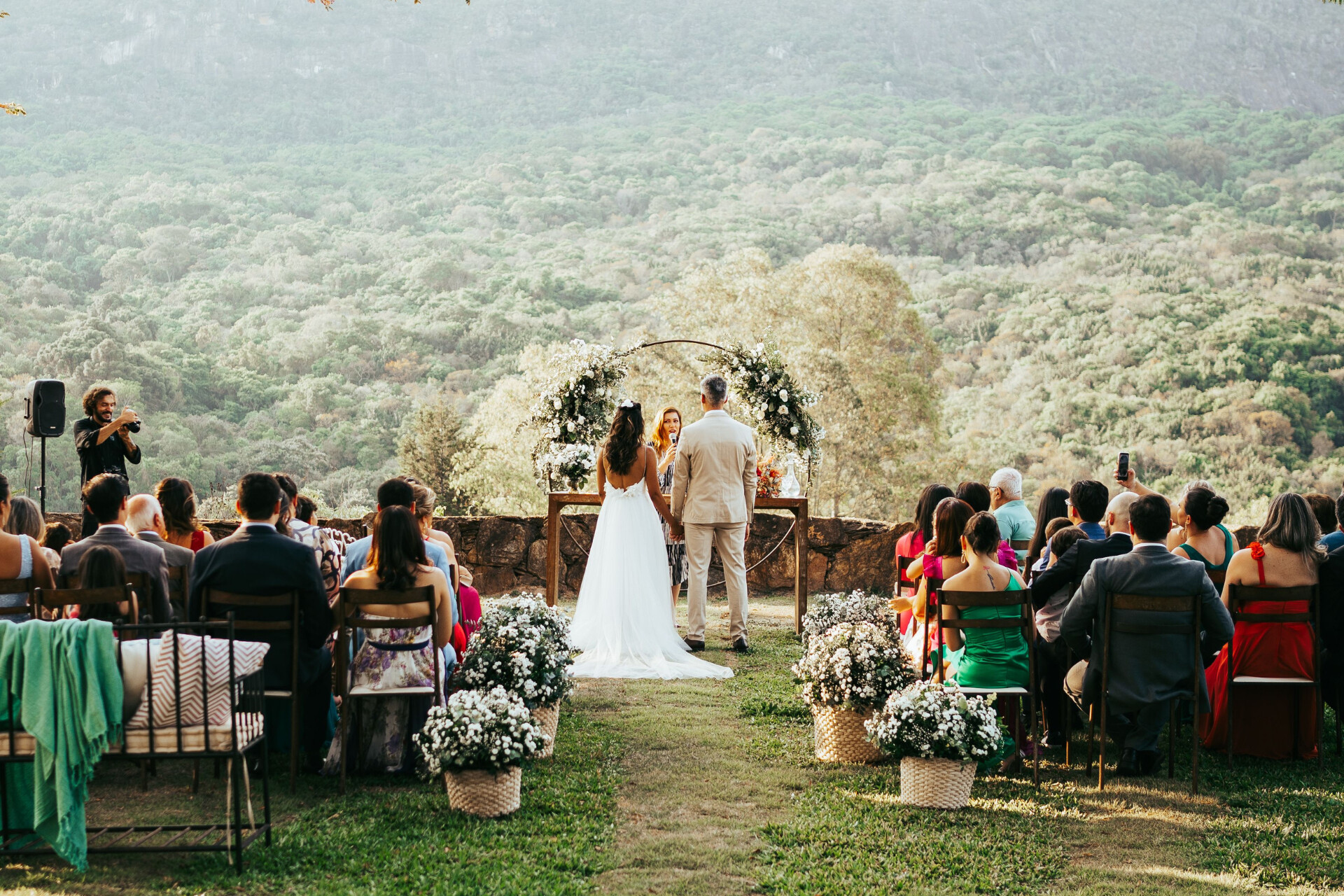 Outdoor wedding ceremony with guests seated on a lawn overlooking a scenic countryside landscape