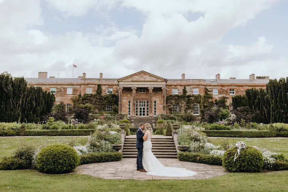 Bride and groom holding hands in the gardens of Hillsborough Castle. The palace wedding venue is in the background.