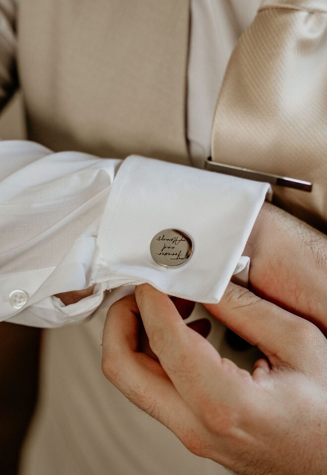 Groom fastening a personalised cufflink on a white shirt, wearing a cream tie and waistcoat.