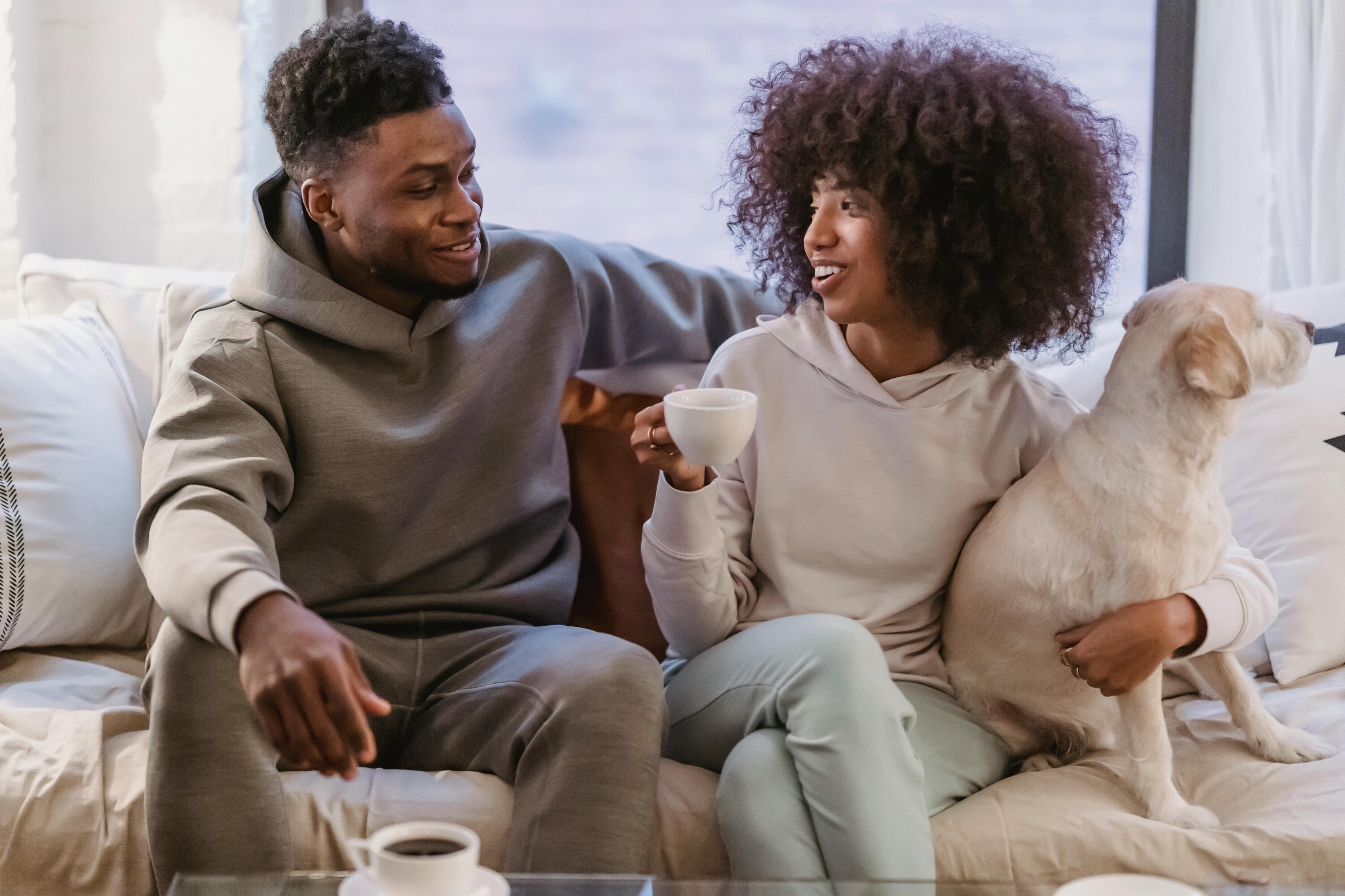 Couple relaxing together on a sofa at home with their dog while drinking coffee and talking.