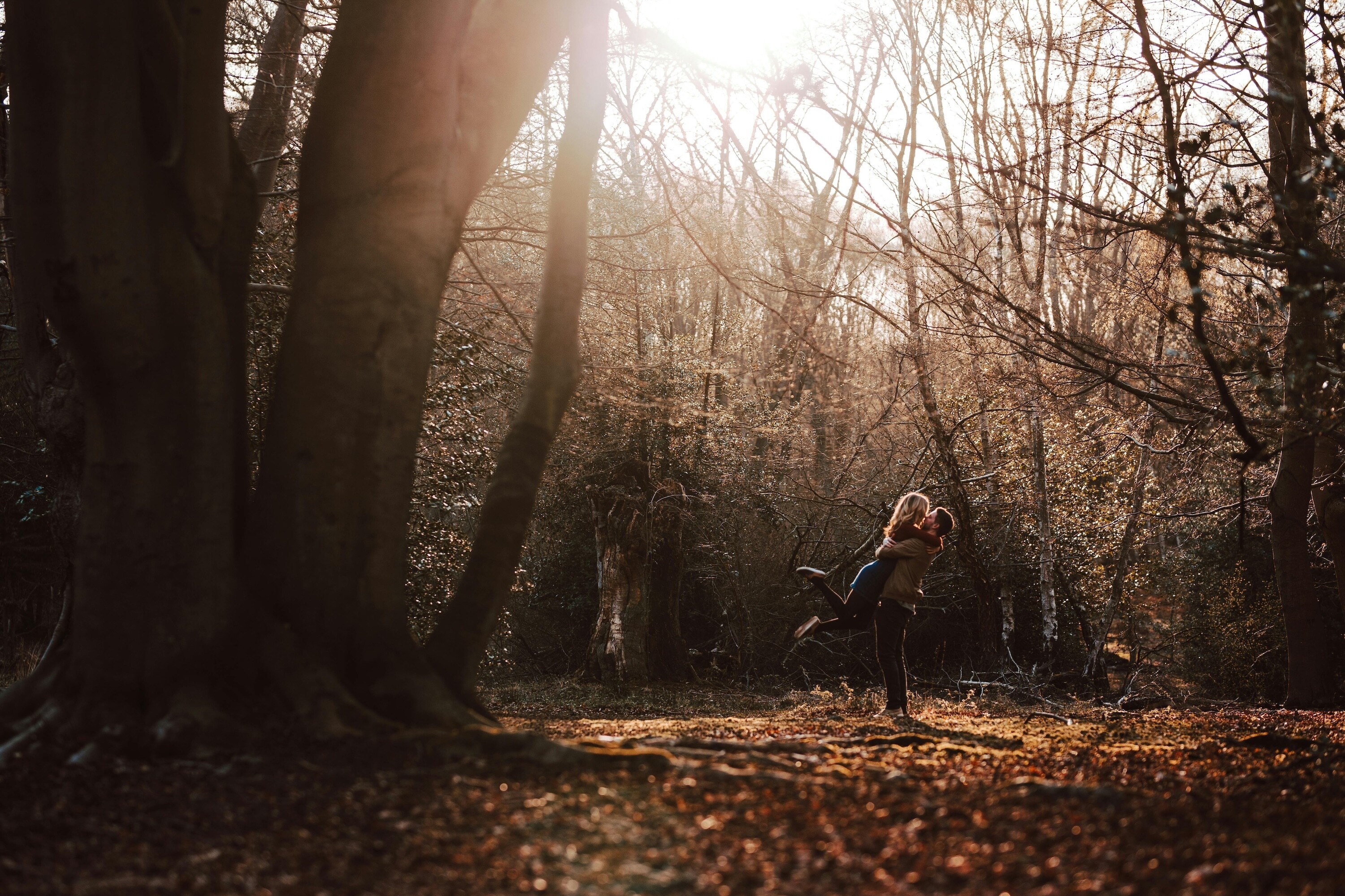 Groom lifting the bride during their engagement photoshoot in the woods, surrounded by fallen leaves.