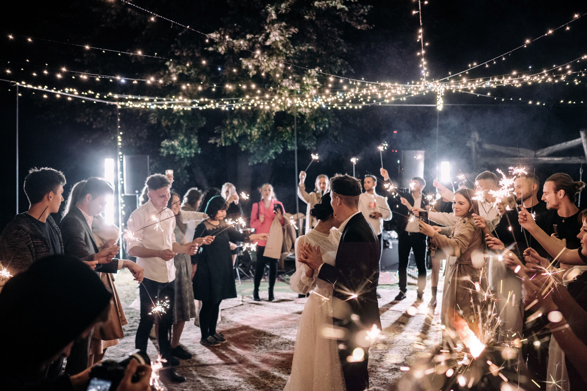 Newlyweds sharing a kiss surrounded by guests holding sparklers during an evening wedding celebration.