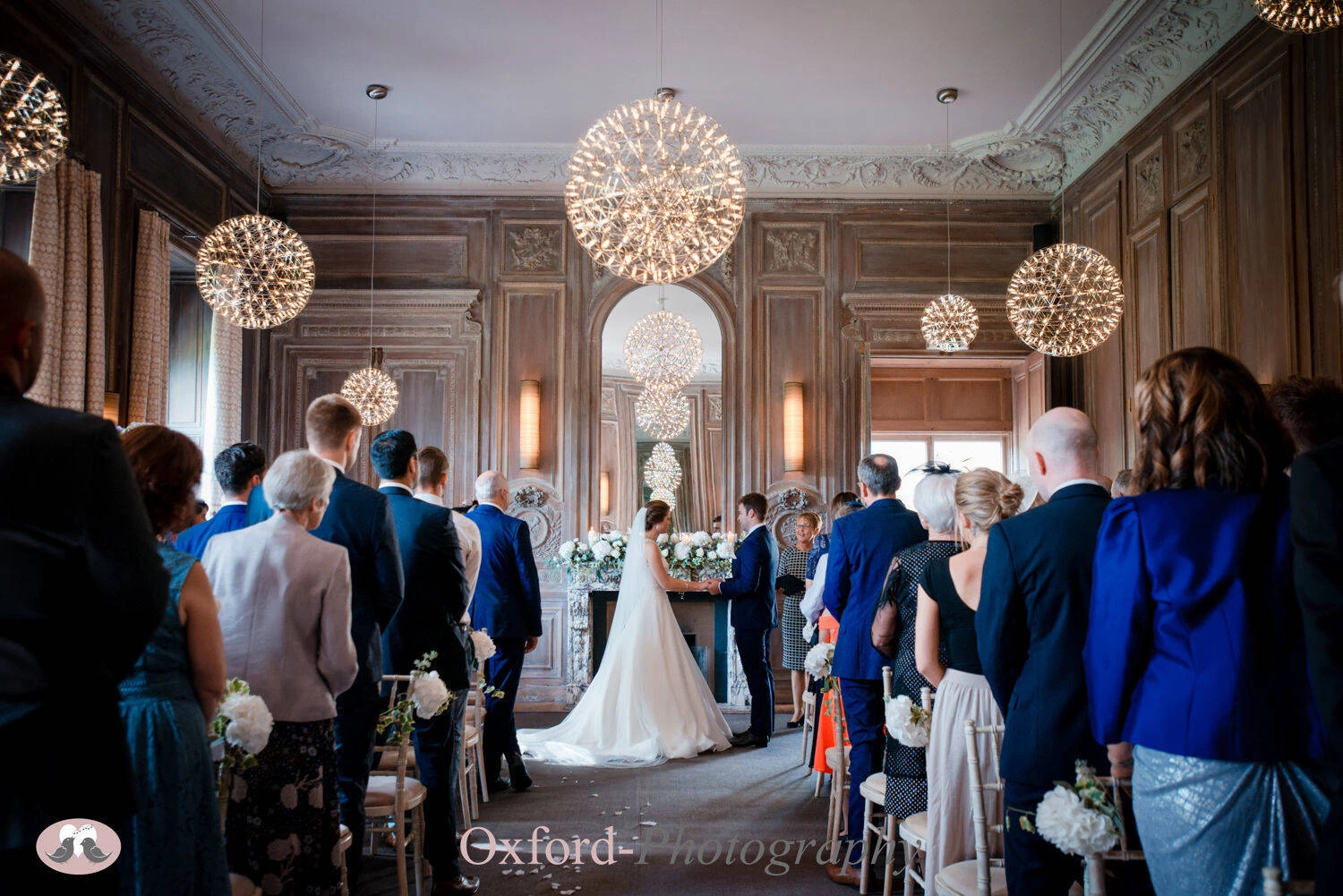 Wedding ceremony in wood panelled room, with couple stood in front of fireplace. Captured by Aimee Kirkham Oxford-Photography