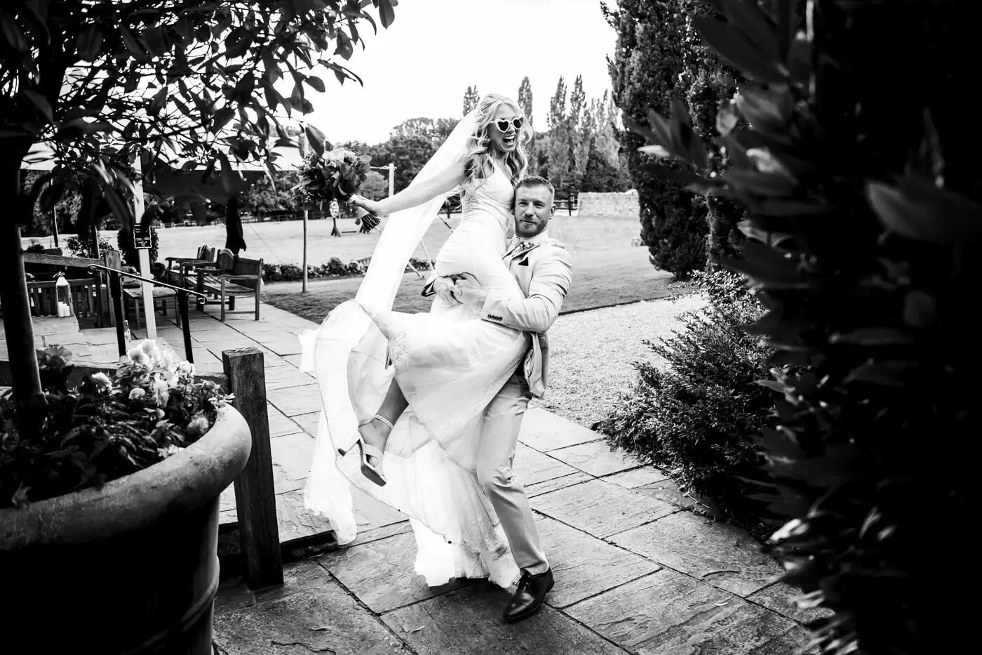 Black and white photo of a groom lifting the bride outside, as she wears white heart-shaped sunglasses. Taken by Marcus Charter Photography in Buckinghamshire.