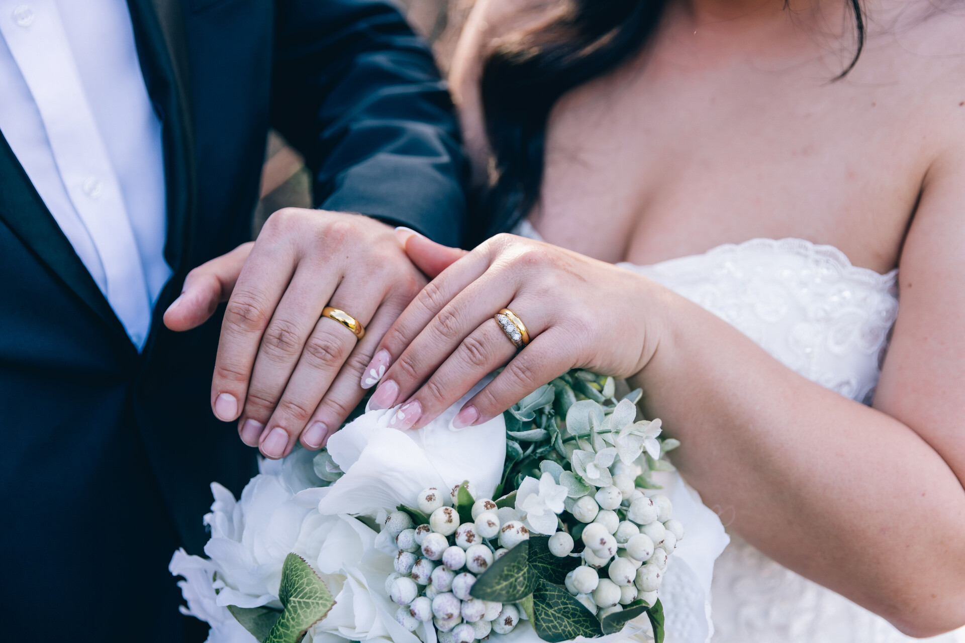Close-up of a bride and groom showing their wedding rings above a white floral bouquet