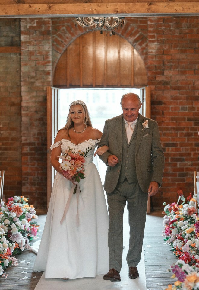 Father of the bride walks daughter down the aisle with faux flowers framing