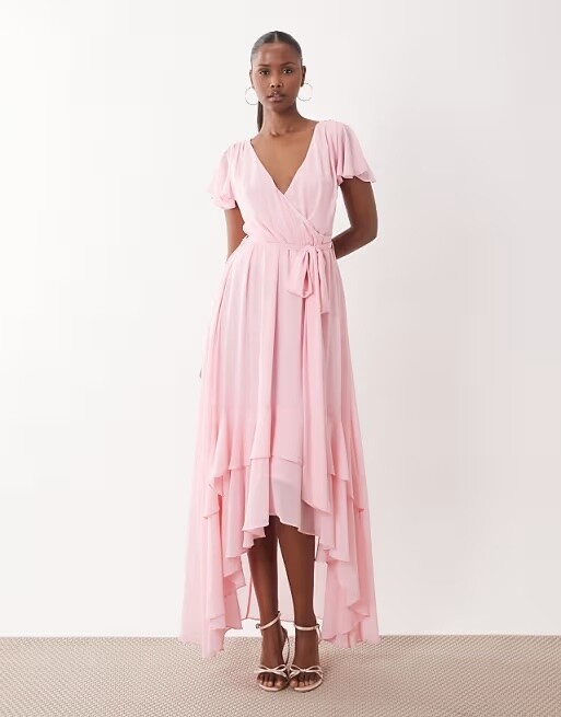 Model wearing a blush pink chiffon bridesmaid dress with flutter sleeves, wrap bodice, tie waist and high-low hem.