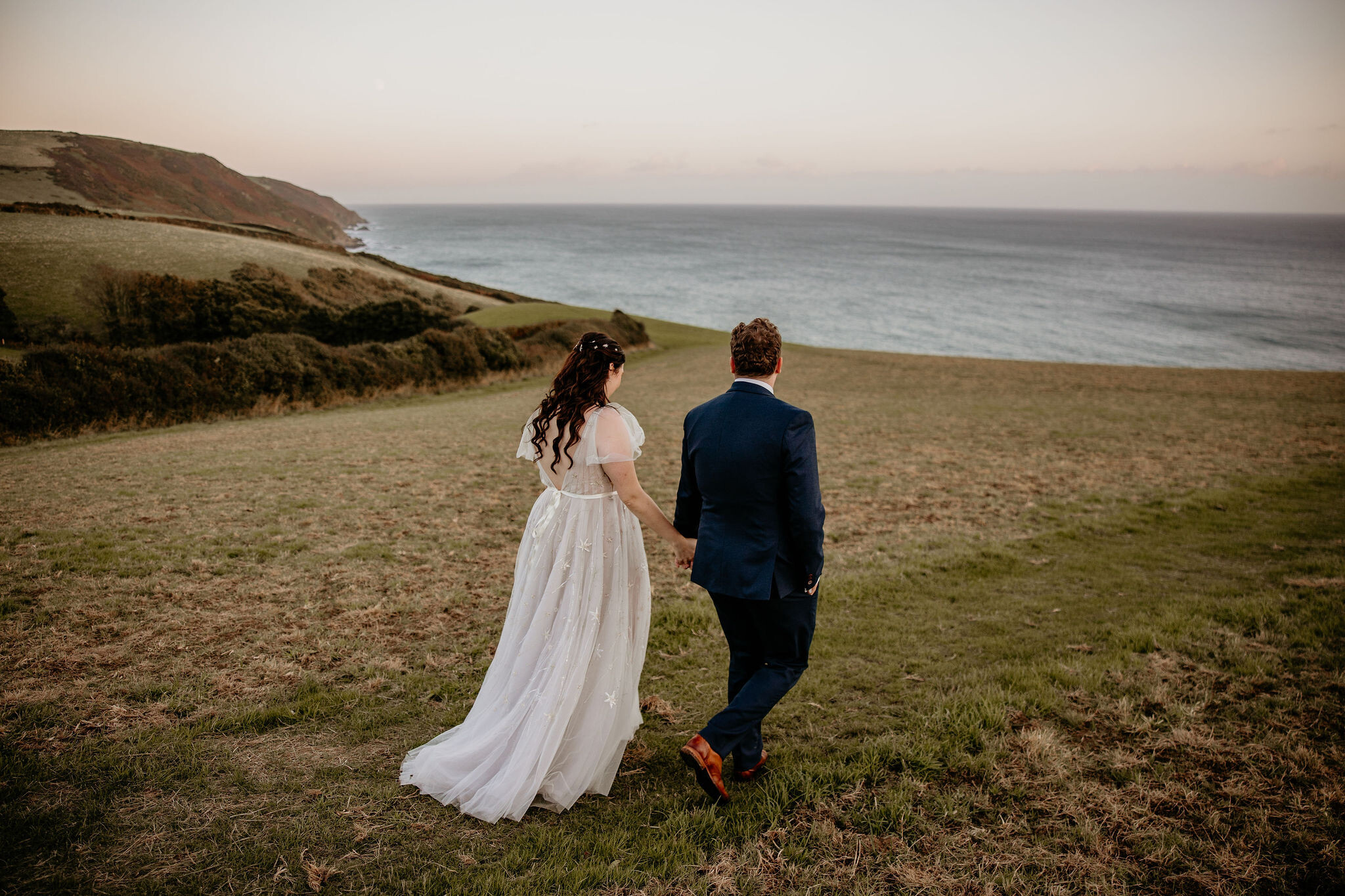 Bride and Groom walk across sea facing cliff in Cornwall