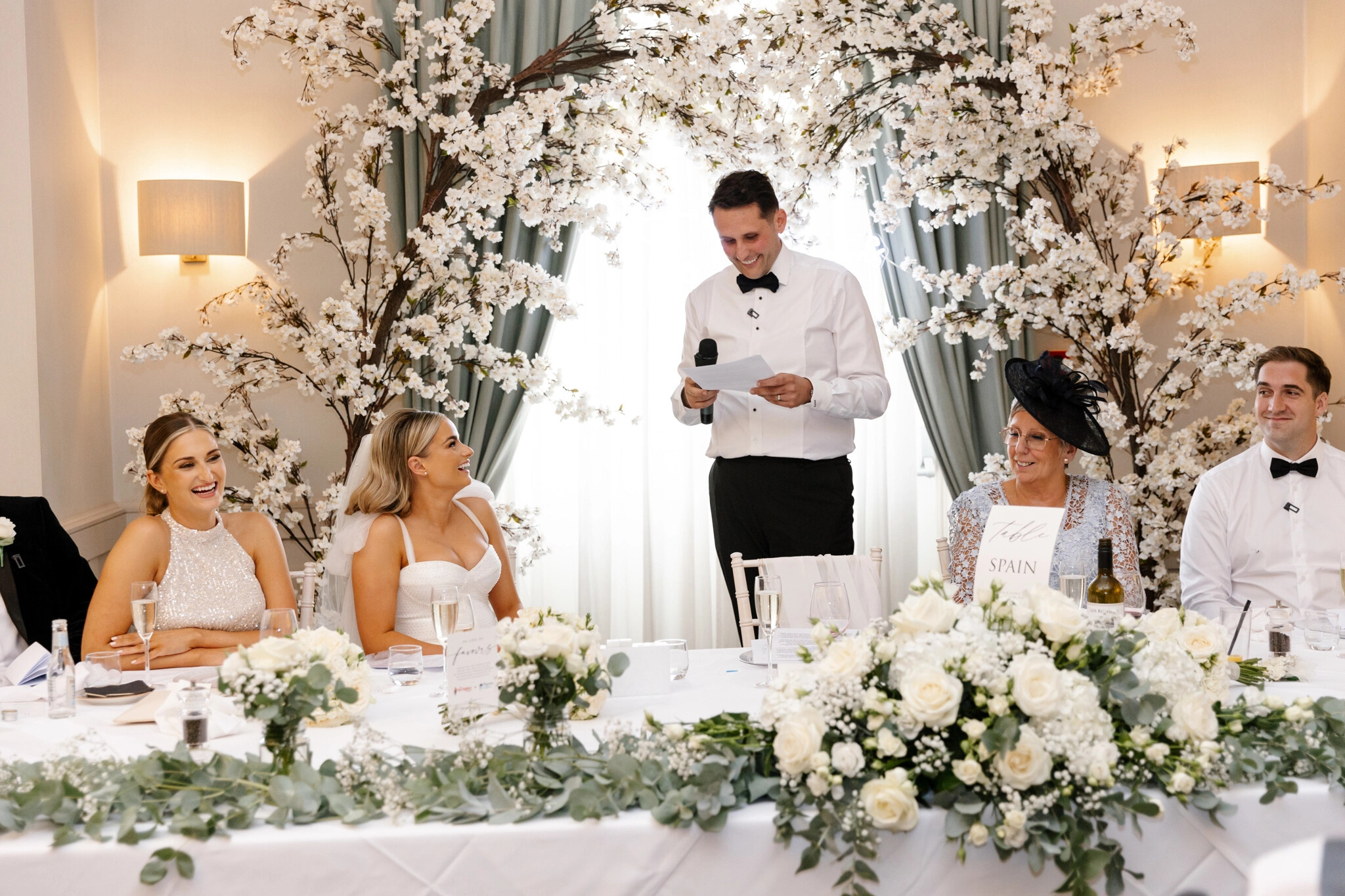 Groom stands framed by floral arrangements giving a speech behind the top table. The bride smiles at him.