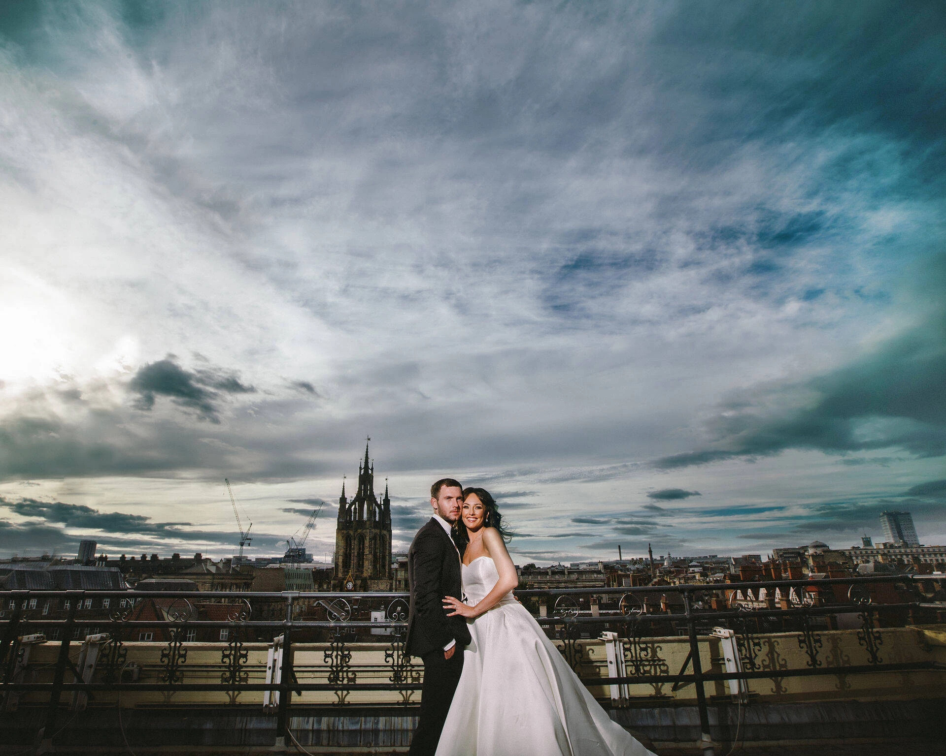 Bride and groom posing on the rooftop terrace at The Vermont Hotel in Newcastle upon Tyne, with the city skyline and cathedral spire under the evening sky.