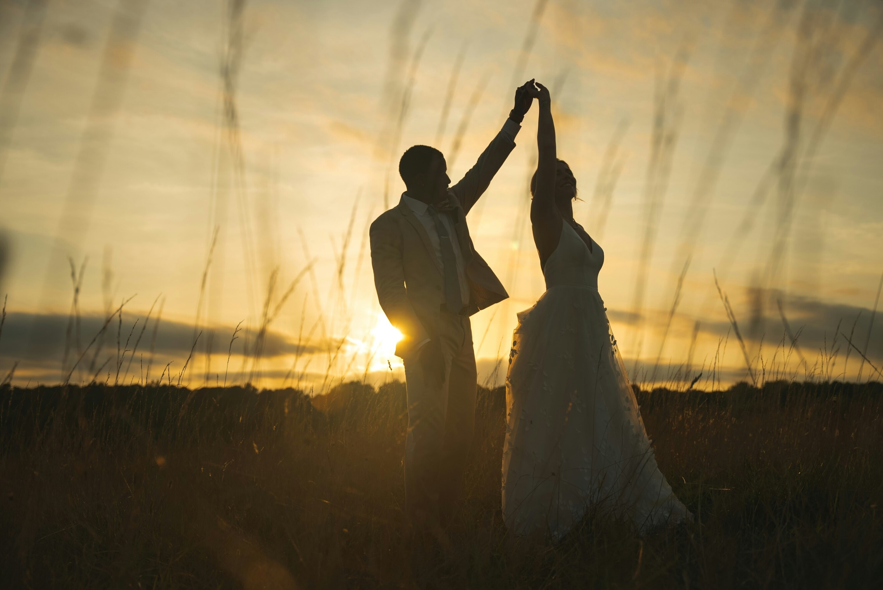 Fine art wedding photography showing a couple silhouetted at sunset in long grass, captured with soft light.