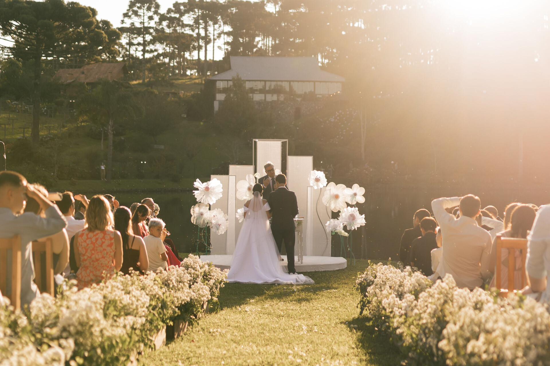 Elegant outdoor wedding ceremony with a flower-lined aisle and guests gathered beside a tranquil lakeside setting.