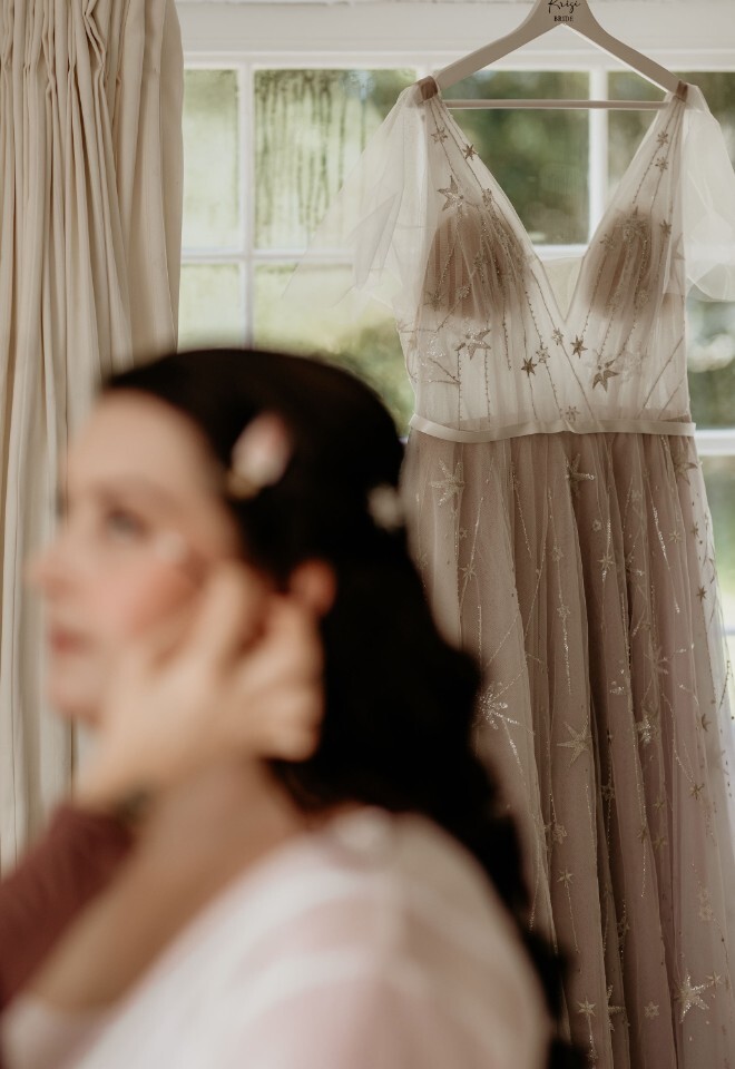 Bride’s wedding dress hanging, featuring sheer fabric with star embroidery and a soft blush lining, with a personalised hanger reading “Krisi Bride" whilst bride gets ready