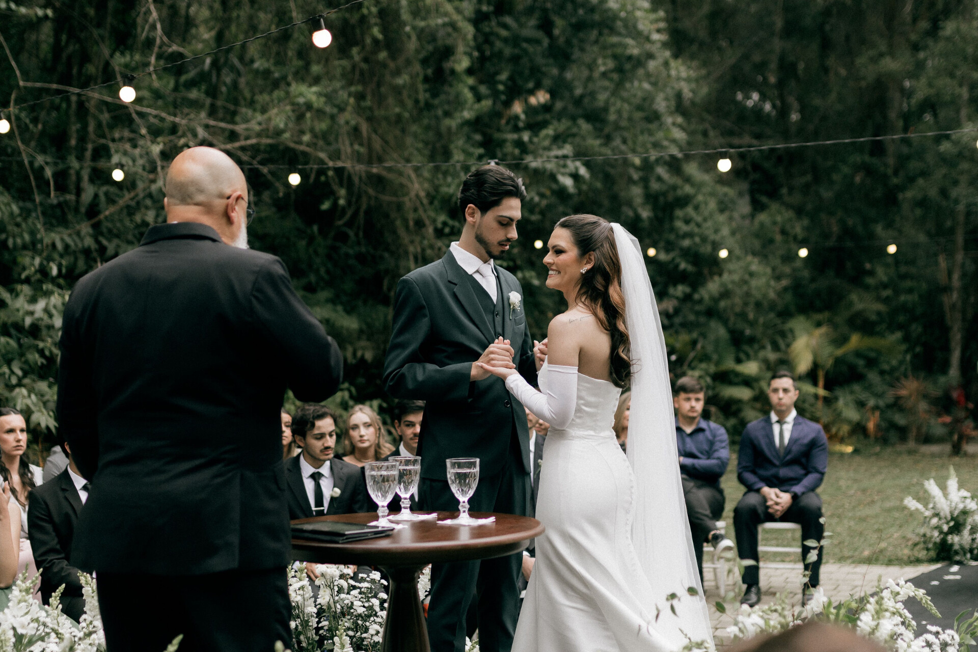 Couple exchanging vows during an intimate outdoor woodland wedding ceremony with guests seated beneath festoon lights.