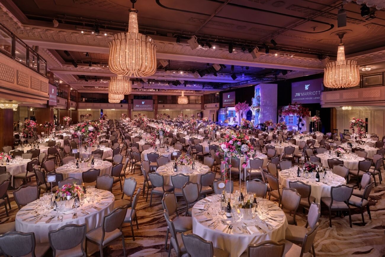 The Great Room at Grosvenor House set for a wedding reception with round dining tables, floral centrepieces, chandeliers and a large stage.