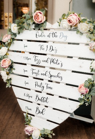 Details of timings for a wedding day on white heart shaped board with flowers at Southdowns Manor West Sussex