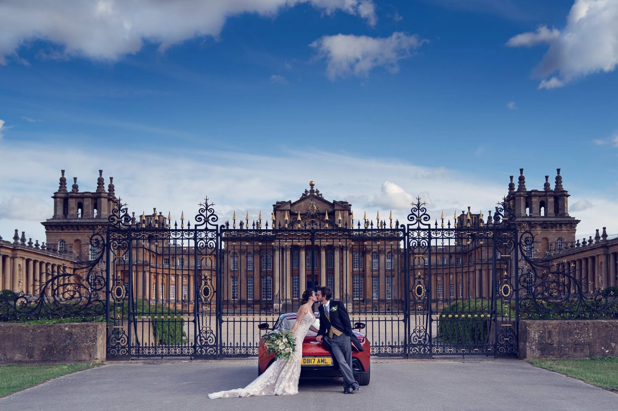 Couple kiss next to car in front of iron gates of Blenheim Palace. The impressive facade of the palace venue is behind the gates.