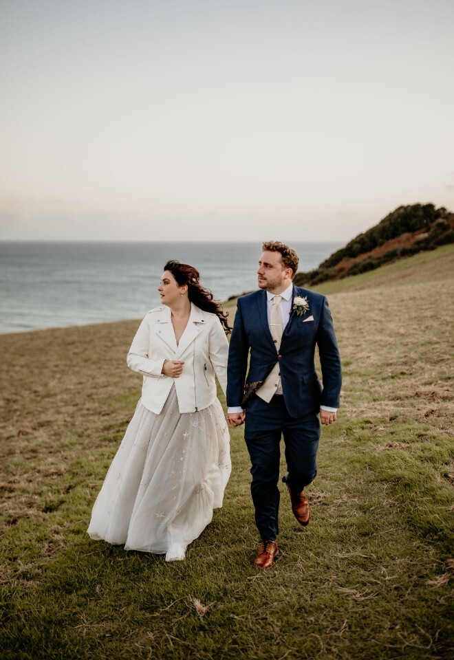 Couple walking along a coastal hillside in wedding attire, holding hands.