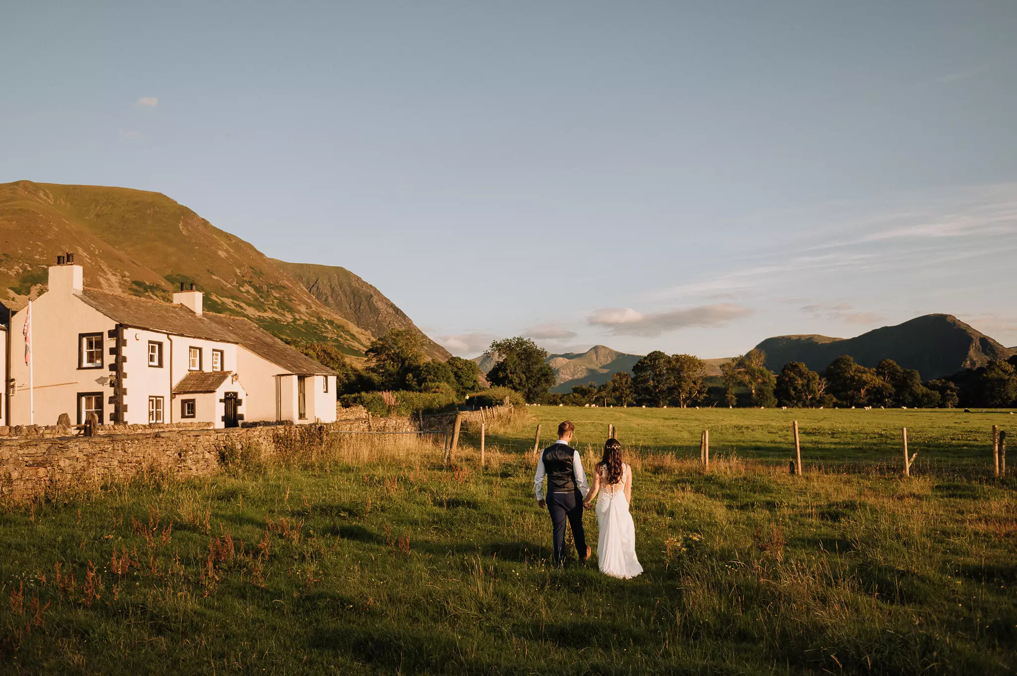 Bride and groom walking hand in hand through fields at New House Farm, with farmhouse and mountain views in warm evening light.