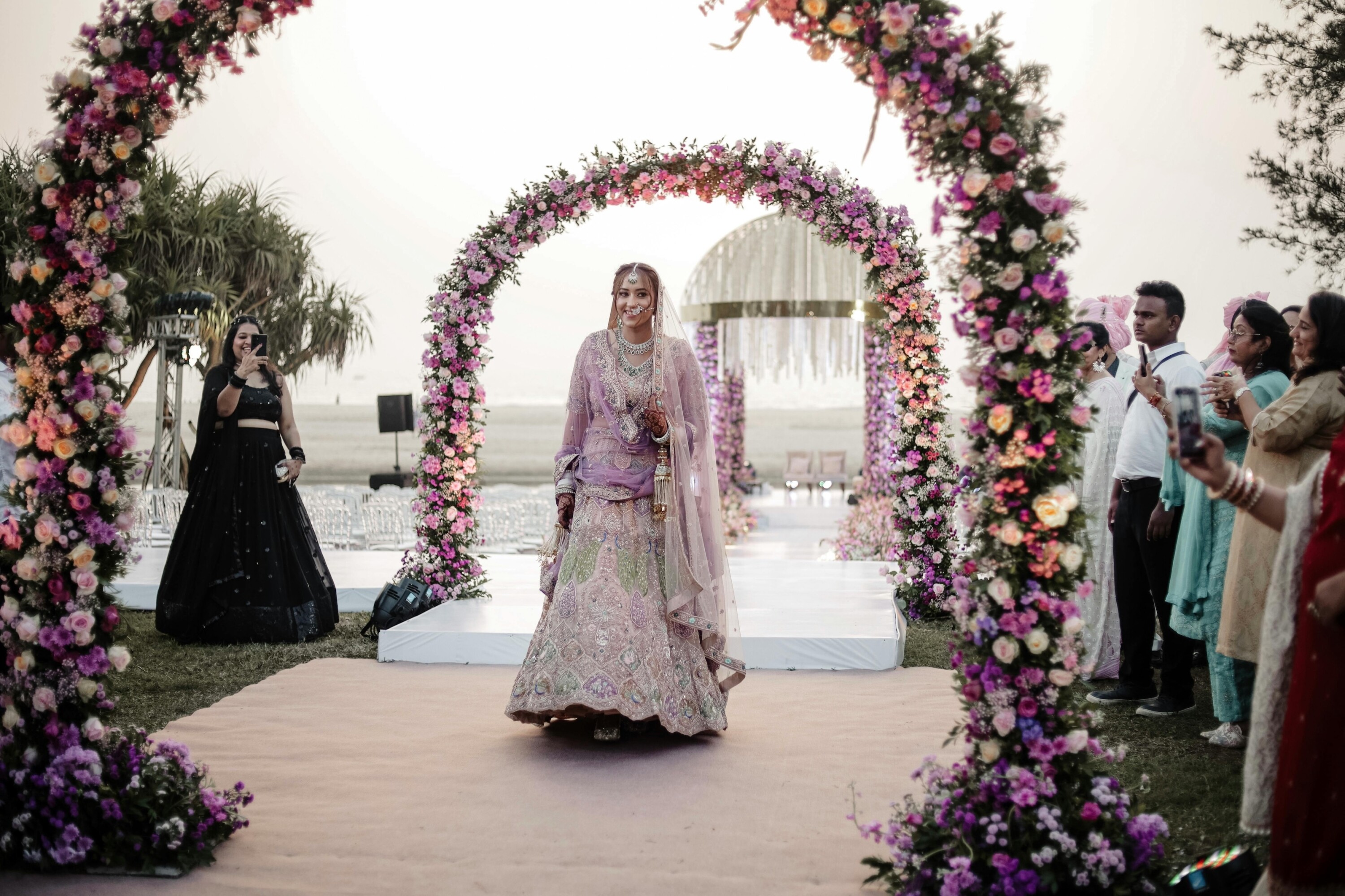 Bride wearing a pink lehenga walks beneath large wedding flower arch arrangements