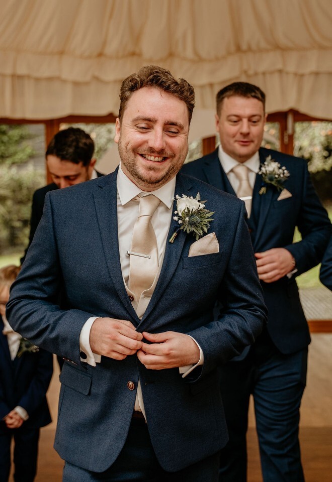 Groom and groomsmen in navy suits getting ready together, adjusting jackets in a light-filled room.