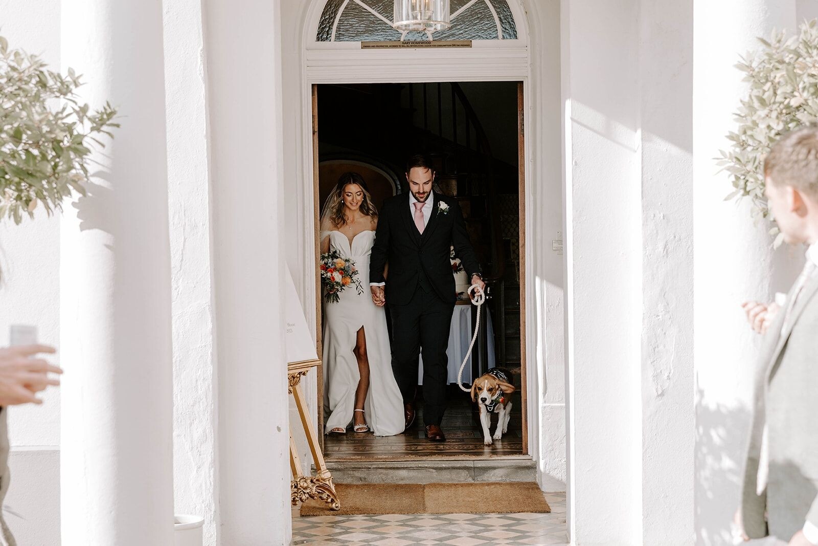 Bride and groom walking out of Glewstone Court with their dog on a lead, holding hands in a bright entranceway.