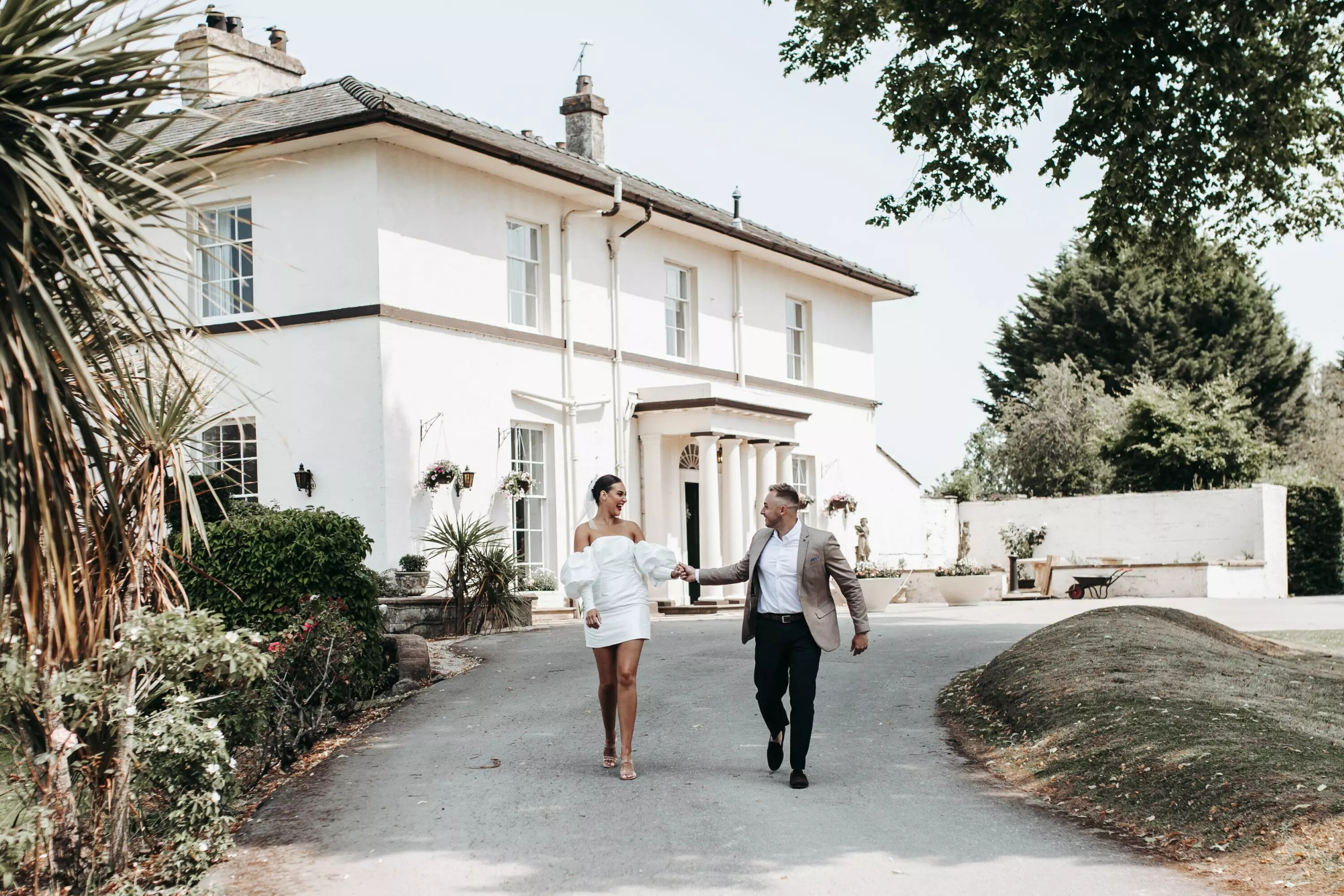 Bride and groom walk hand in hand and smiling outside their wedding venue, Highfield Hall, which is behind them