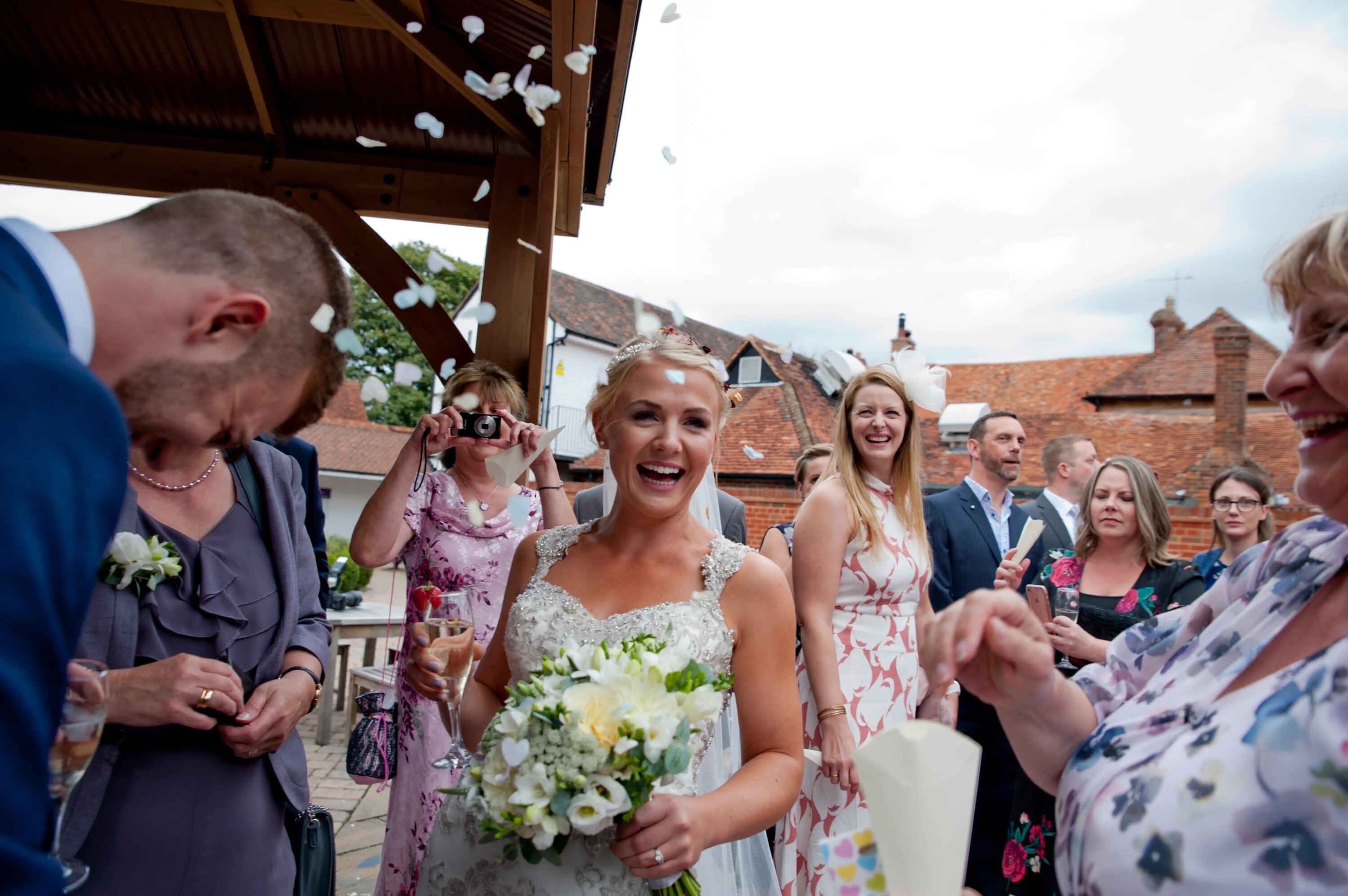 Bride laughing as guests throw confetti outside a rustic venue, holding a white bouquet and champagne glass, surrounded by smiling wedding guests