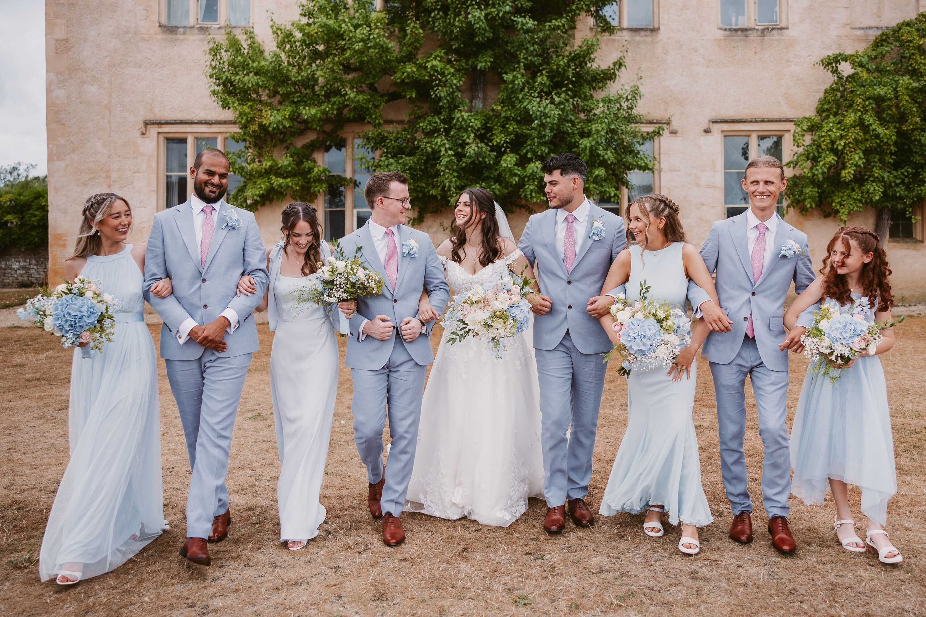Bride and Groom with their wedding party at Cogges Manor Farm wedding venue. Featuring the brides dress, the groom and groomsmen pale blue suits with a light pink tie + the bridesmaids pale blue dress