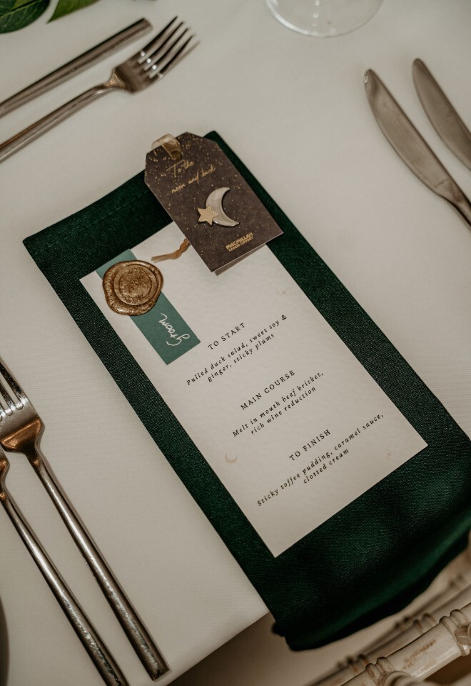 Wedding table place setting with a printed menu, wax seal detail and star and moon place card on a dark green napkin.