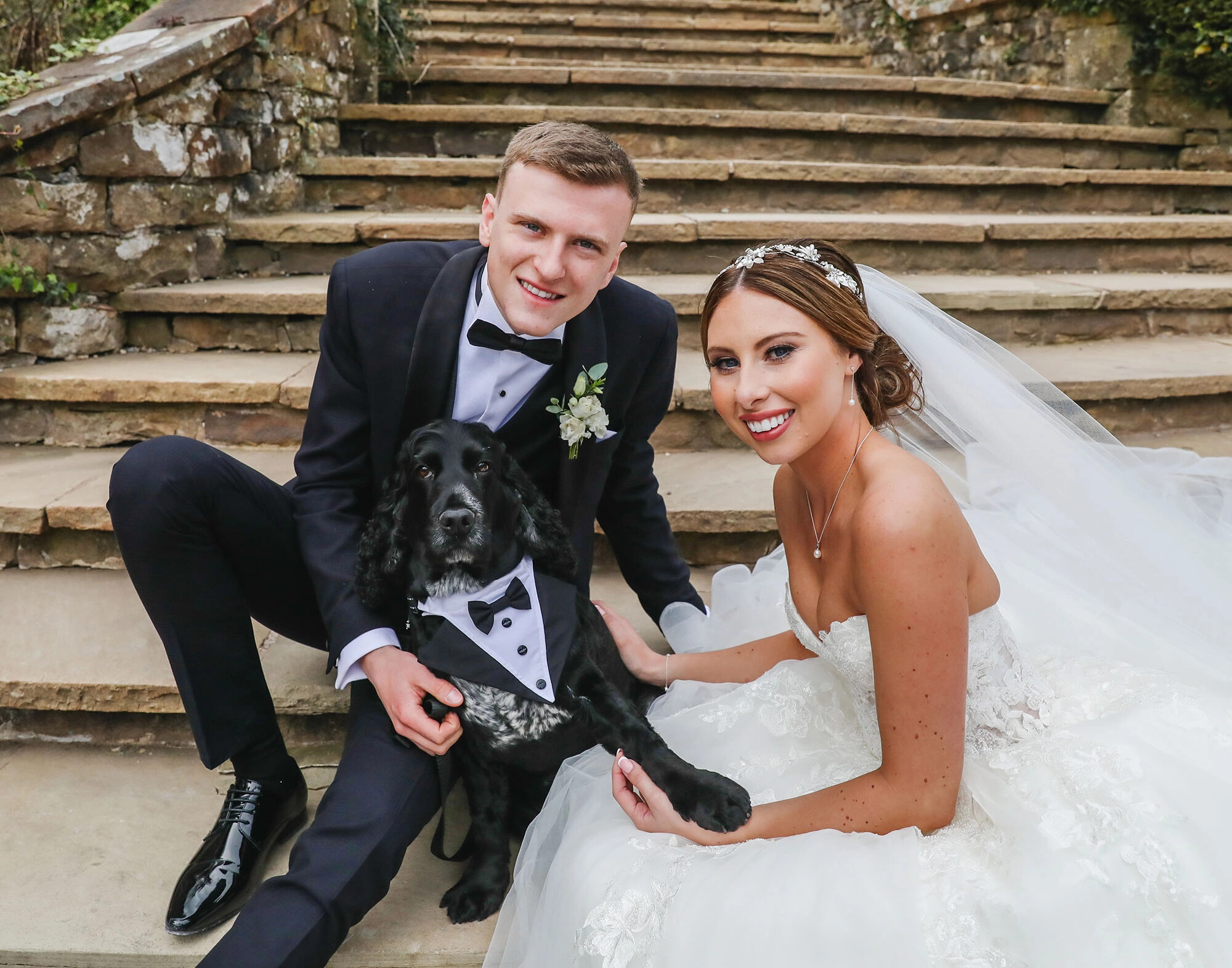 Bride and groom sitting on outdoor steps with their black dog dressed in a tuxedo. Captured by L&L Photography, Lancashire.