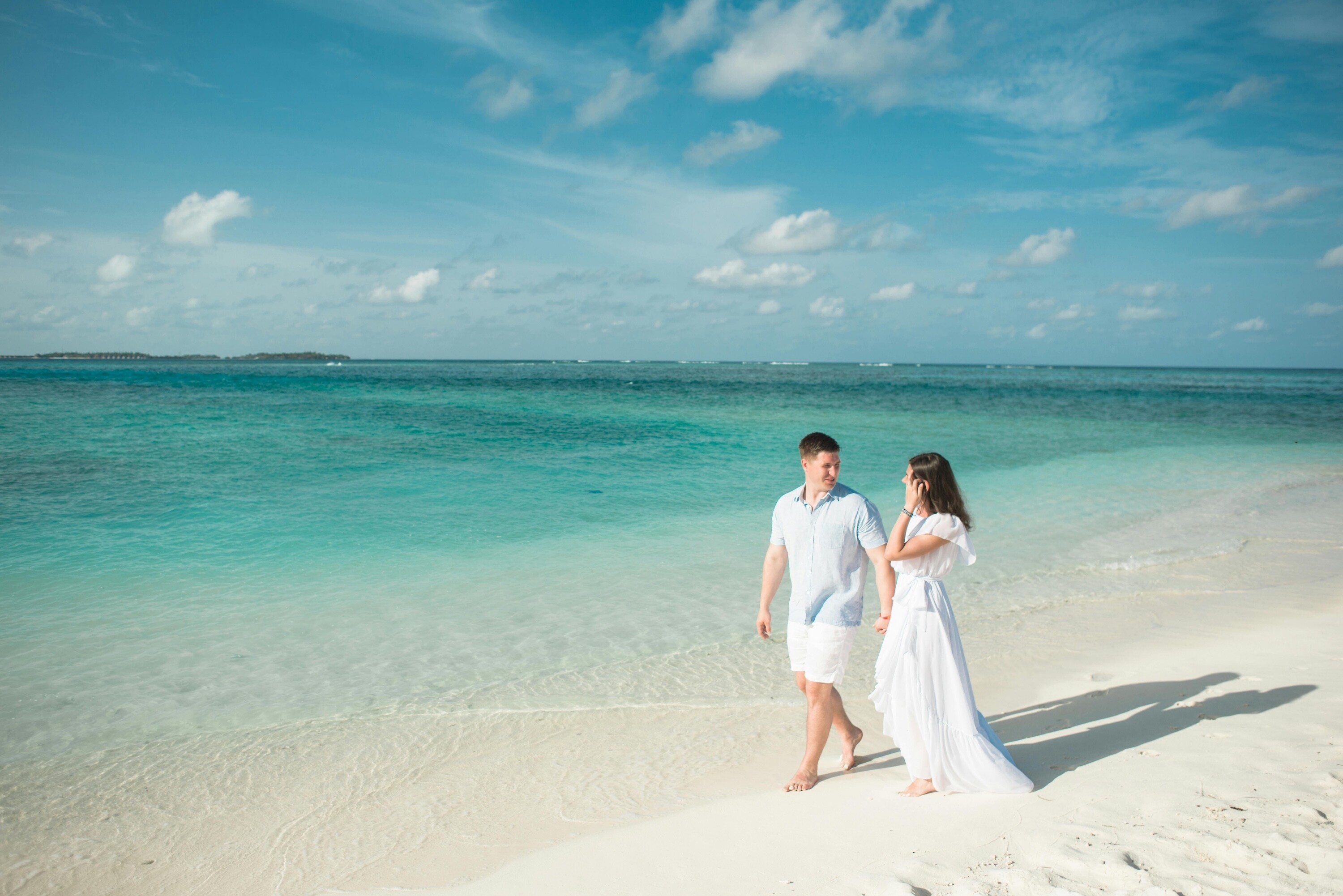 Wedding couple on honeymoon walking hand in hand along a white sand beach in the Maldives, with turquoise sea and blue sky in the background.
