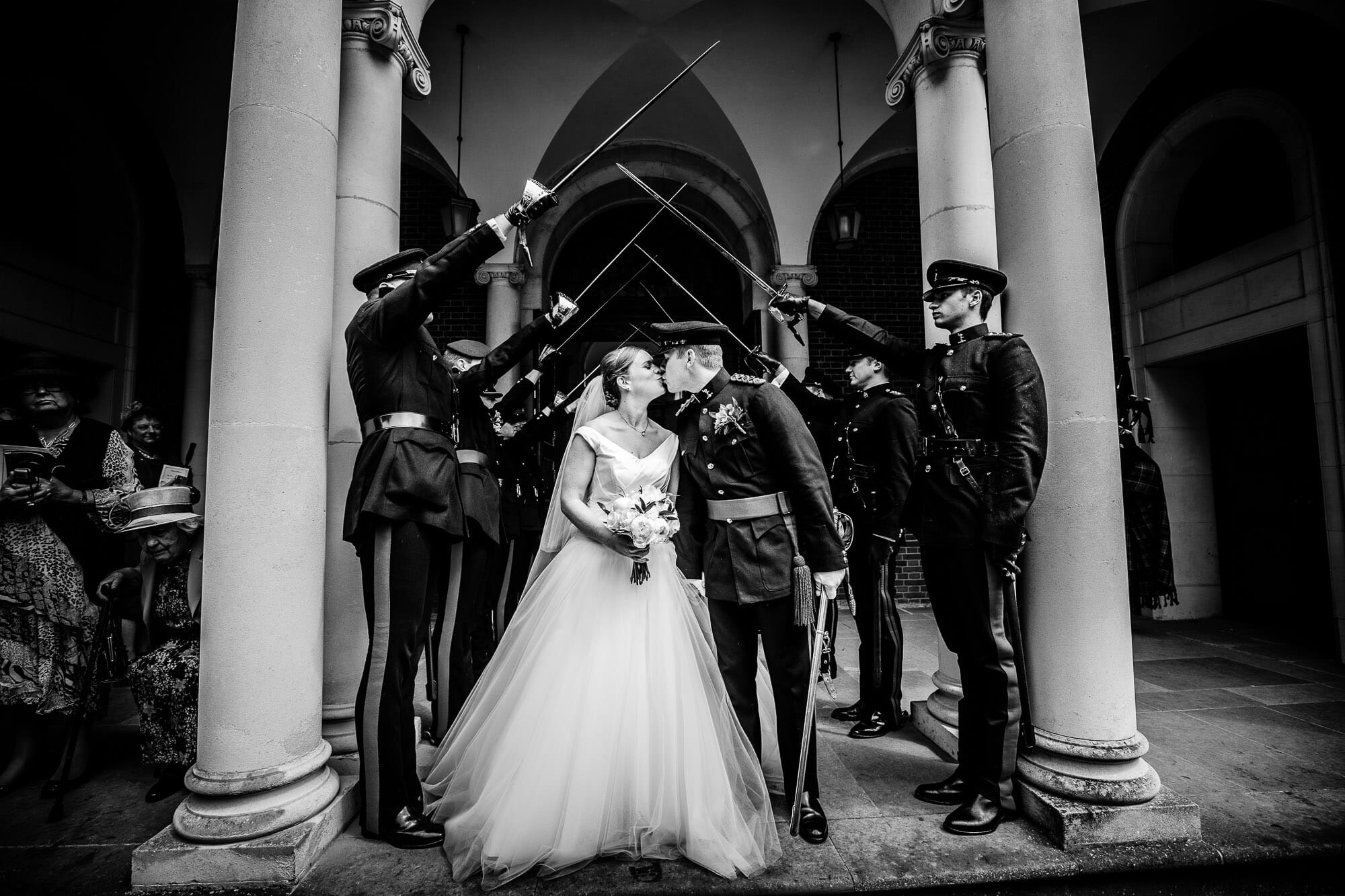 Black and white wedding photo, with the bride and groom under an arch of swords, in between pillars. Taken by Tansley Photography, Hampshire