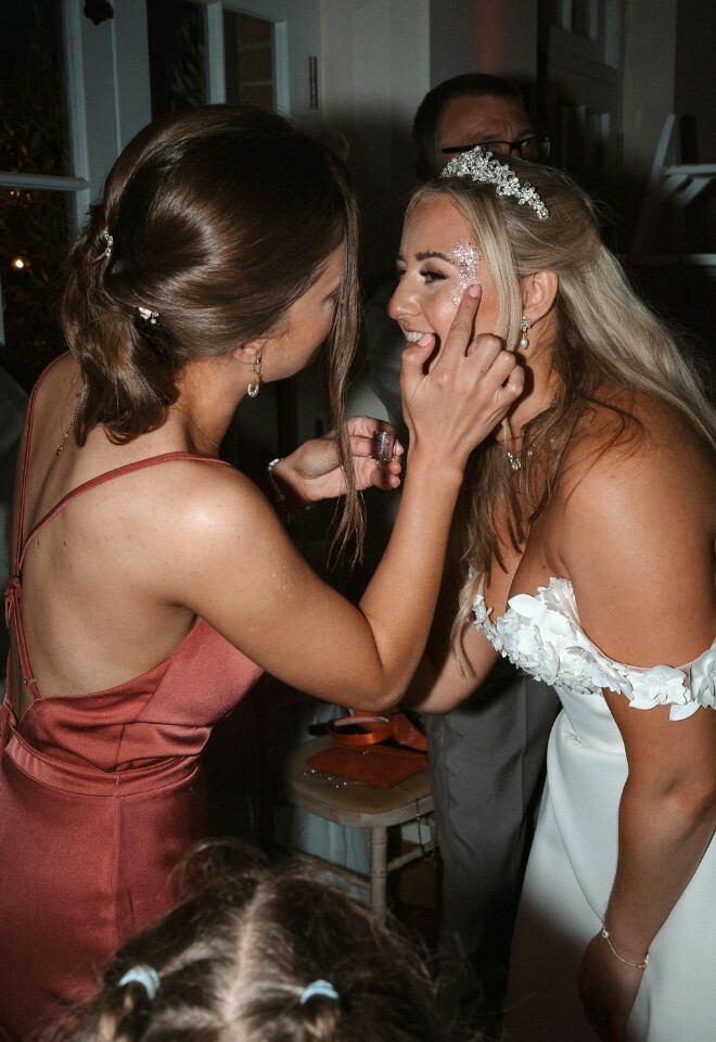 Maid of honour helps bride put glitter on her face at the glitter bar during wedding reception