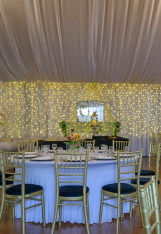 Wedding reception at Duvale Priory in Tiverton, Devon, featuring elegant round tables, gold chairs, a draped ceiling and a backdrop of twinkling fairy lights.