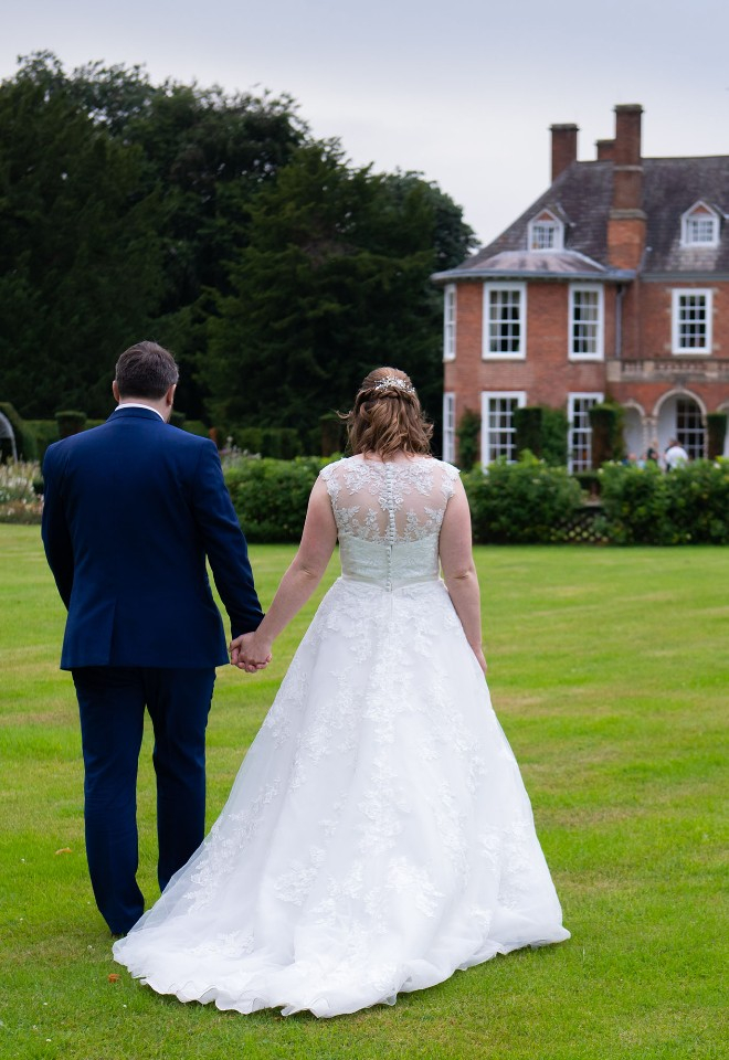 Bride and groom hold hands as they walk away from the camera across the lawns of Sutton Bonington Hall in Leicestershire towards the Hall